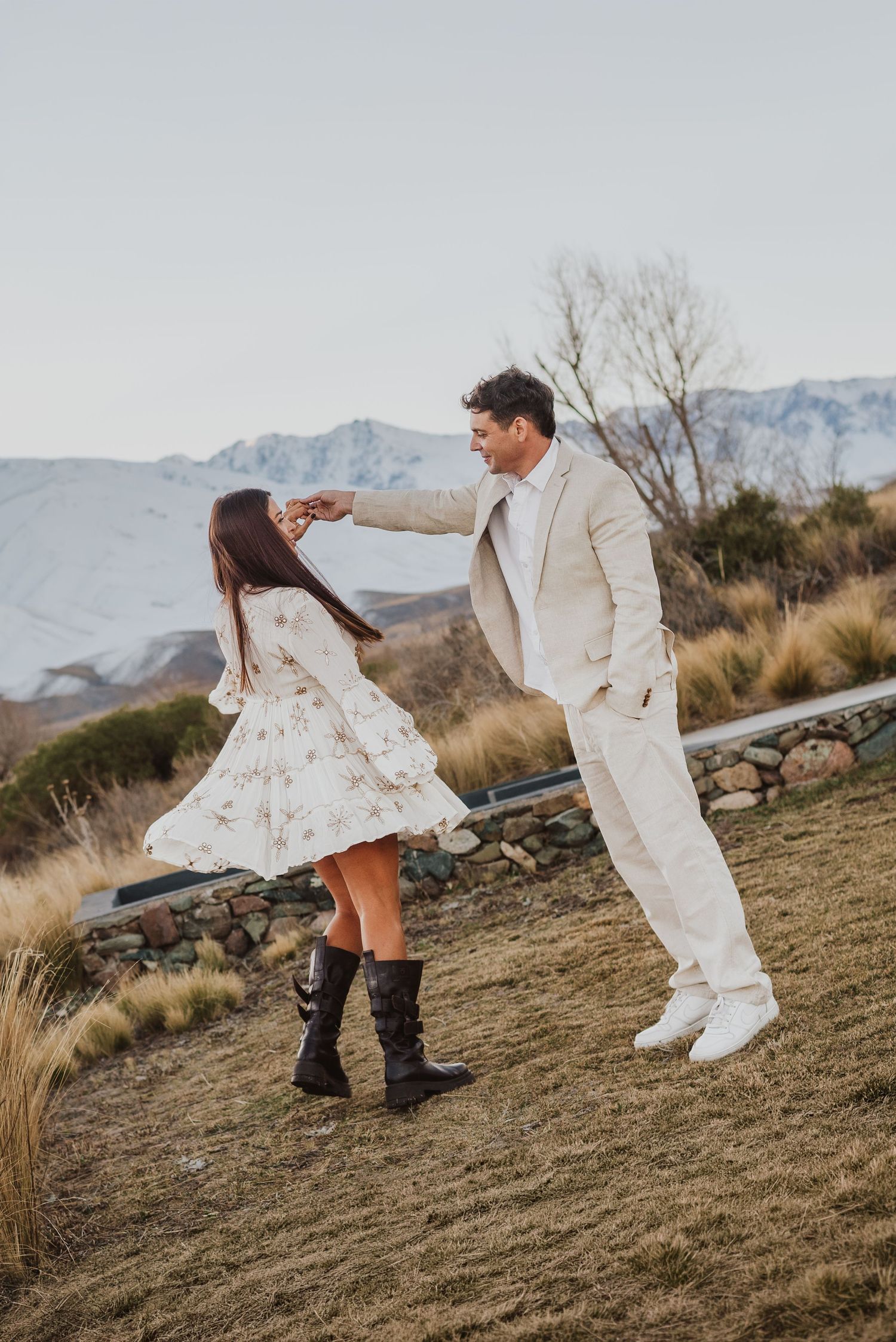 Pareja bailando al aire libre en un paisaje montañoso rústico; la mujer lleva un vestido floral con botas y el hombre un traje color crema.