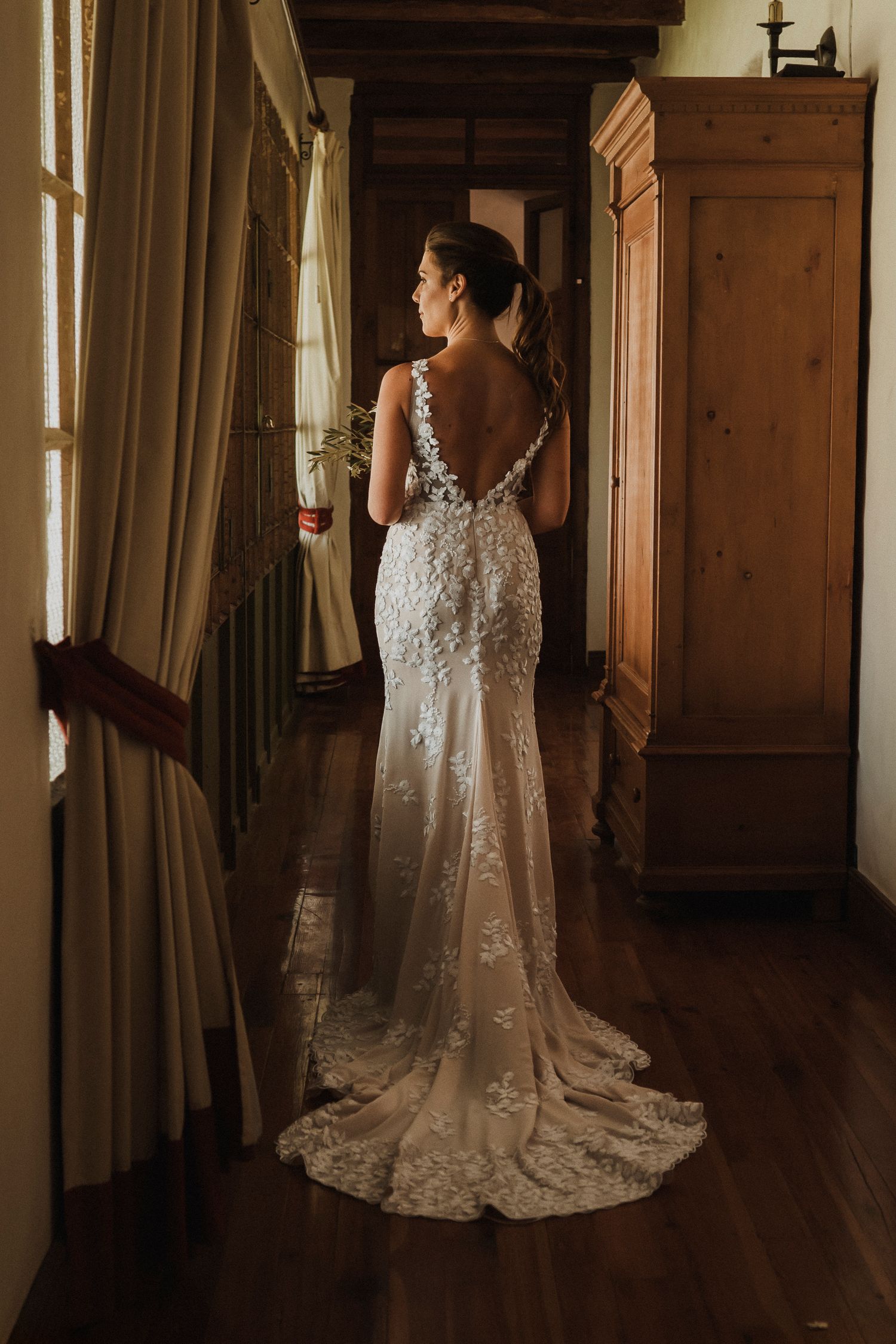 A bride in a white lace wedding dress with a long train stands in an elegant wood-paneled room.
