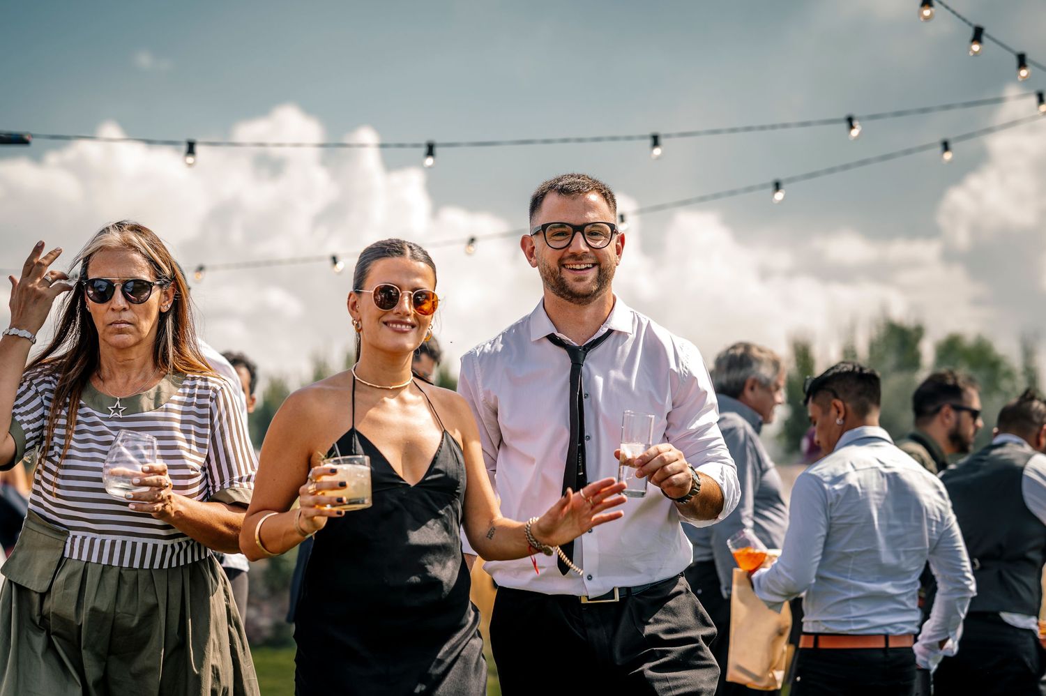 Group of people enjoying drinks and socializing at an outdoor event with string lights overhead.