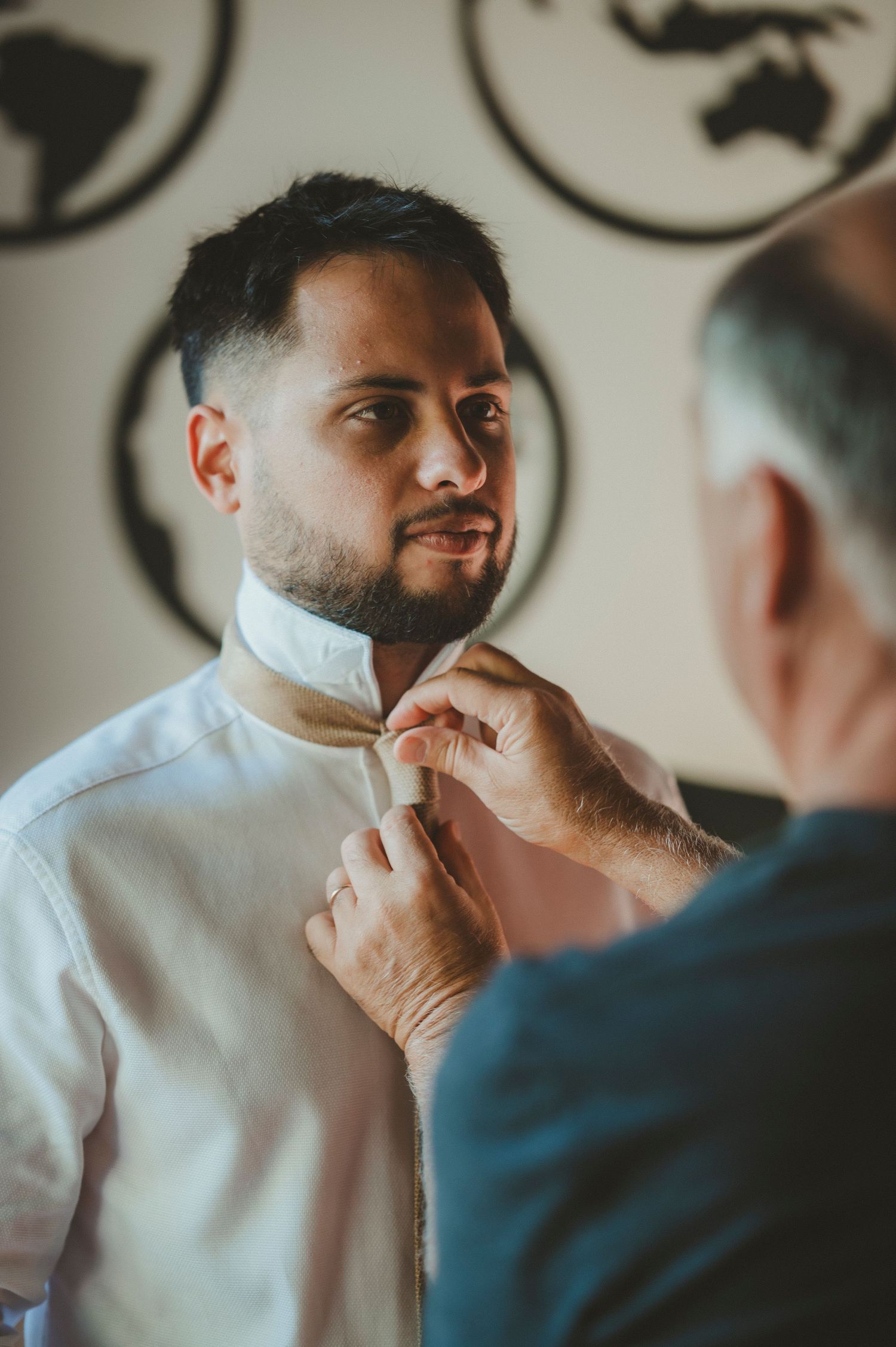 Someone helps adjust a white dress shirt collar in preparation for a formal event, with decorative circular wall art in background.