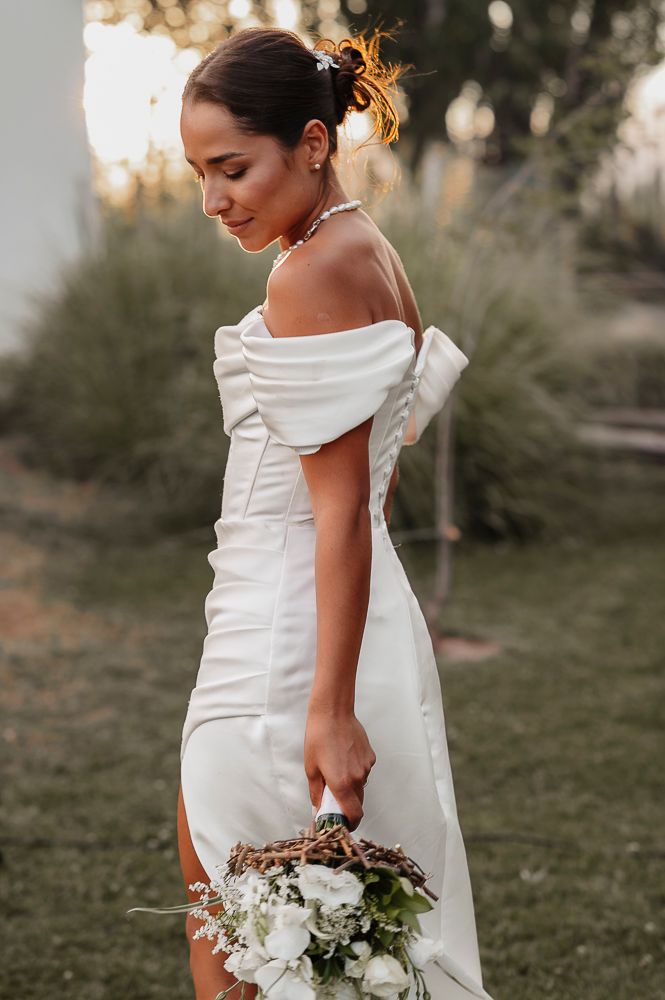 Elegant bride in white off-shoulder wedding dress holds a delicate bouquet of white flowers in outdoor garden setting. wedding in mendoza