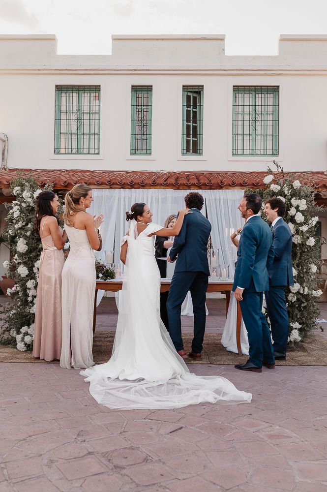 Wedding party gathers on a stone patio with white floral decorations and building facade in the background. wedding in mendoza