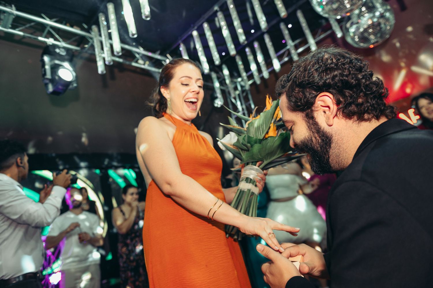 Dancers in orange outfits perform energetic moves at a lively nightclub with colorful lighting.