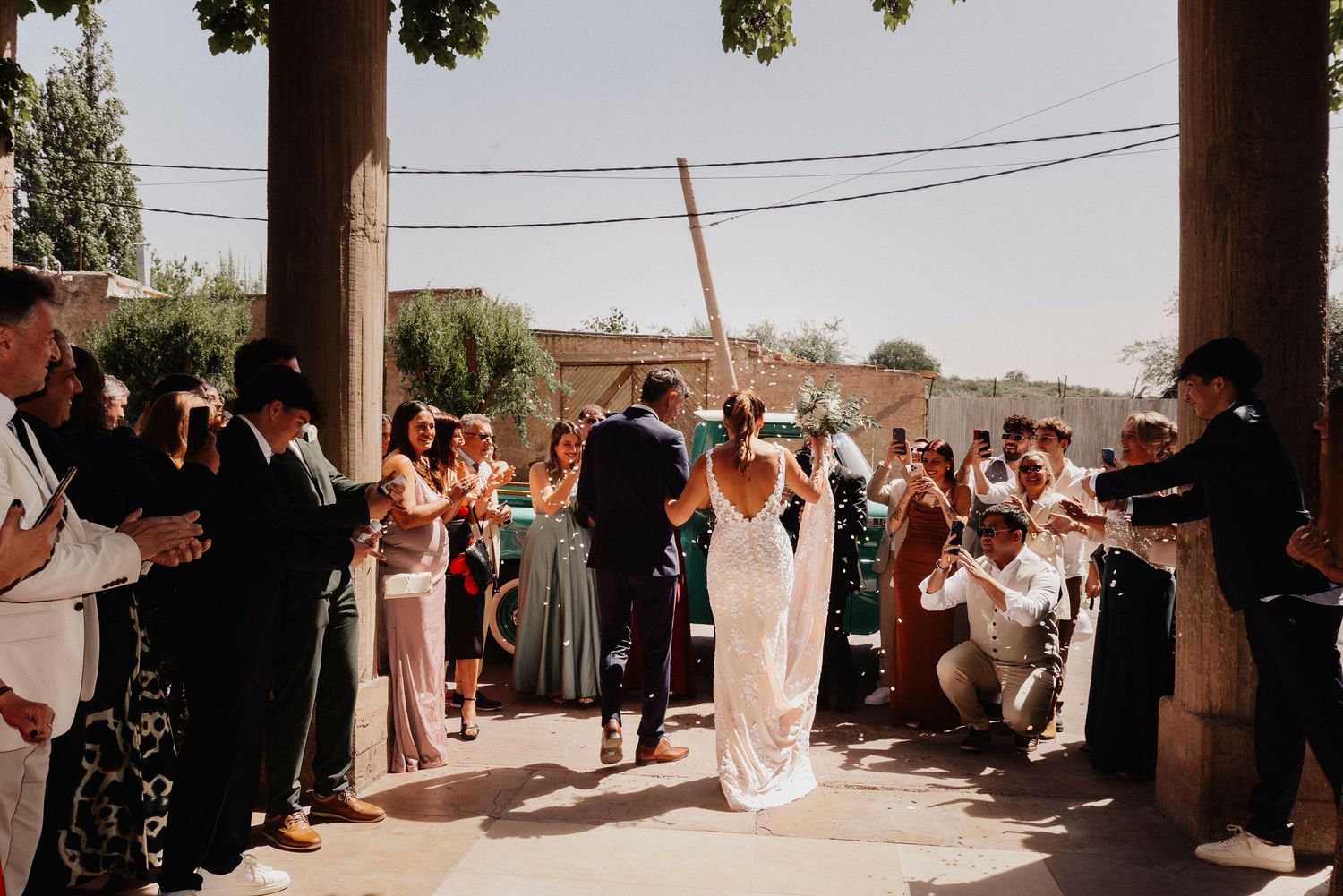 Wedding guests celebrate as newlyweds walk through an outdoor courtyard during a sun-filled ceremony.