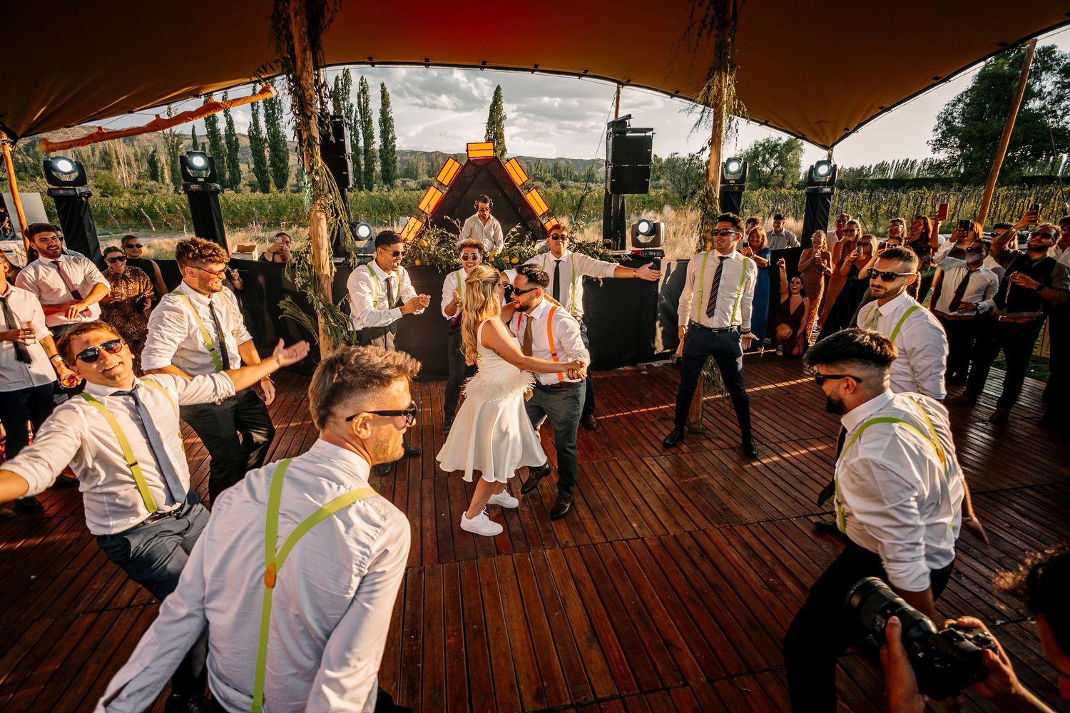 Wedding guests dancing and celebrating on a wooden deck under a tent canopy during an outdoor reception celebration.