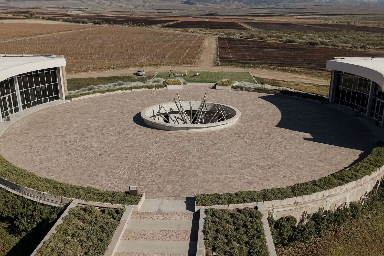 Aerial view of a circular stone plaza with a central sculpture, flanked by modern buildings overlooking vast farmland.