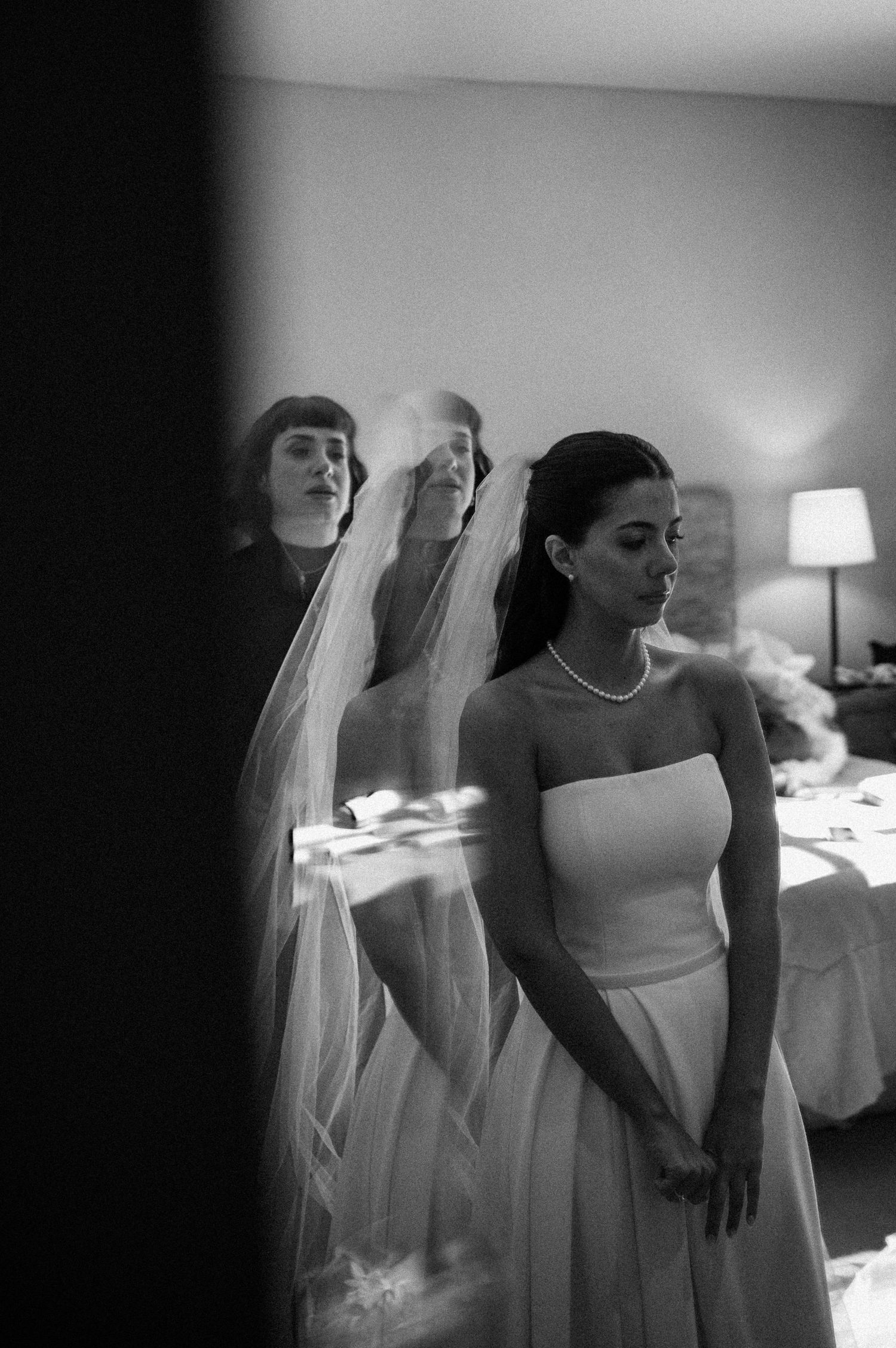 Black and white photo of a bride in strapless gown and veil with pearl necklace, reflected in mirror with bridesmaid.
