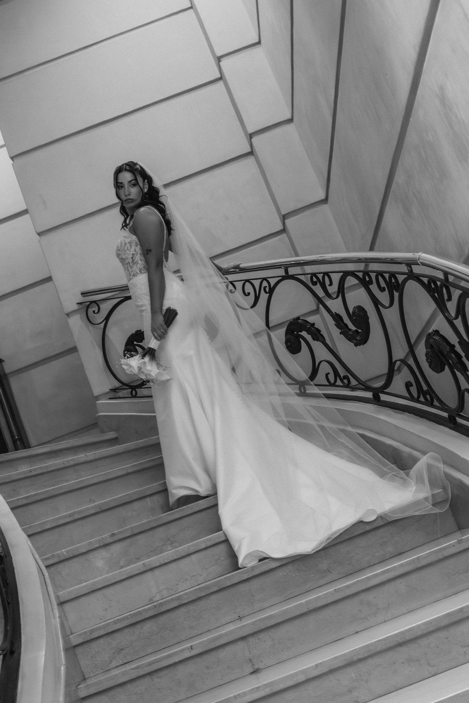 En una fotografía en blanco y negro, se ve a una novia con un vaporoso vestido blanco y velo descendiendo por una elegante escalera curva co