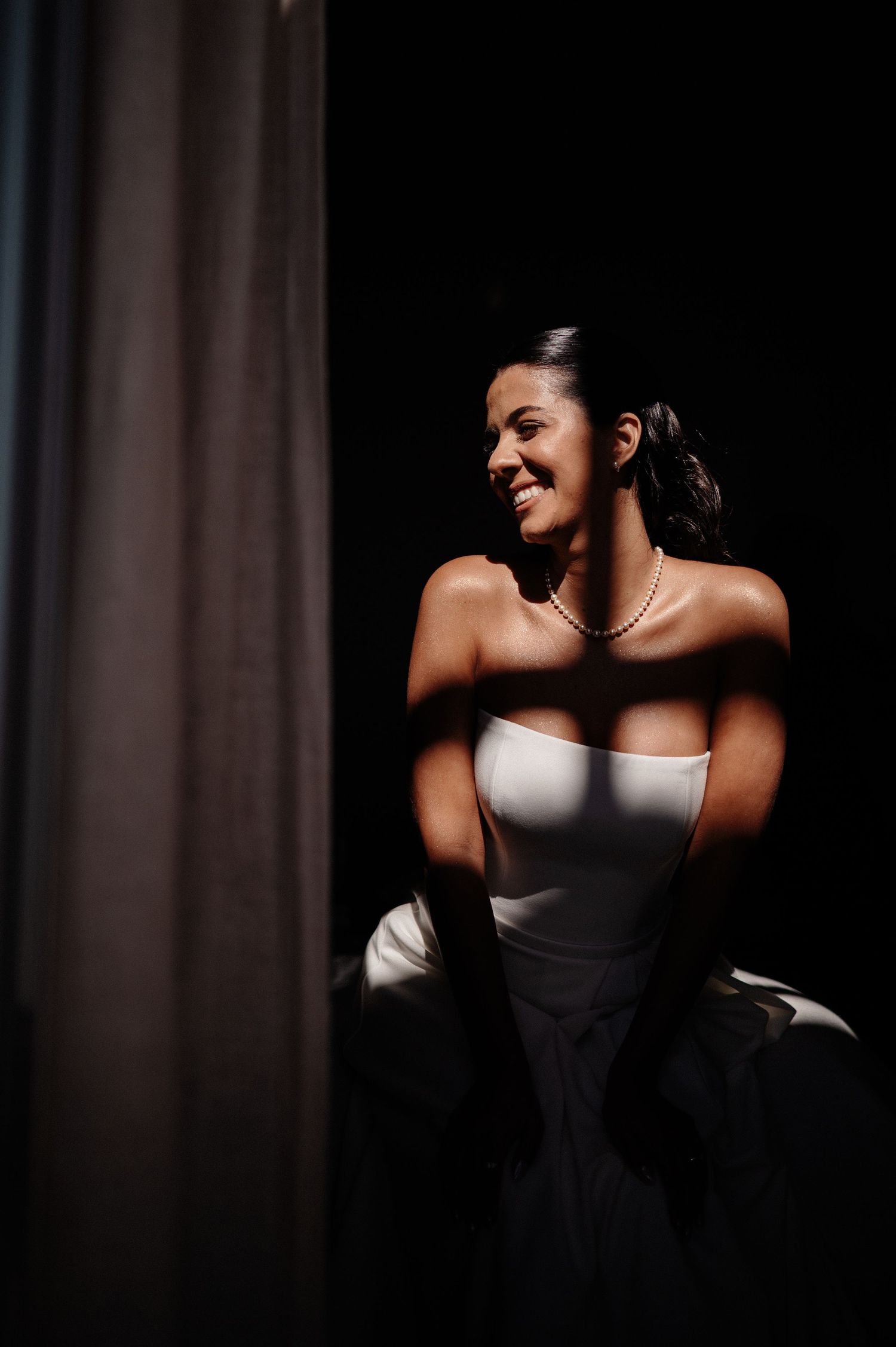 Smiling bride in strapless white gown and pearl necklace, bathed in dramatic light against dark background.