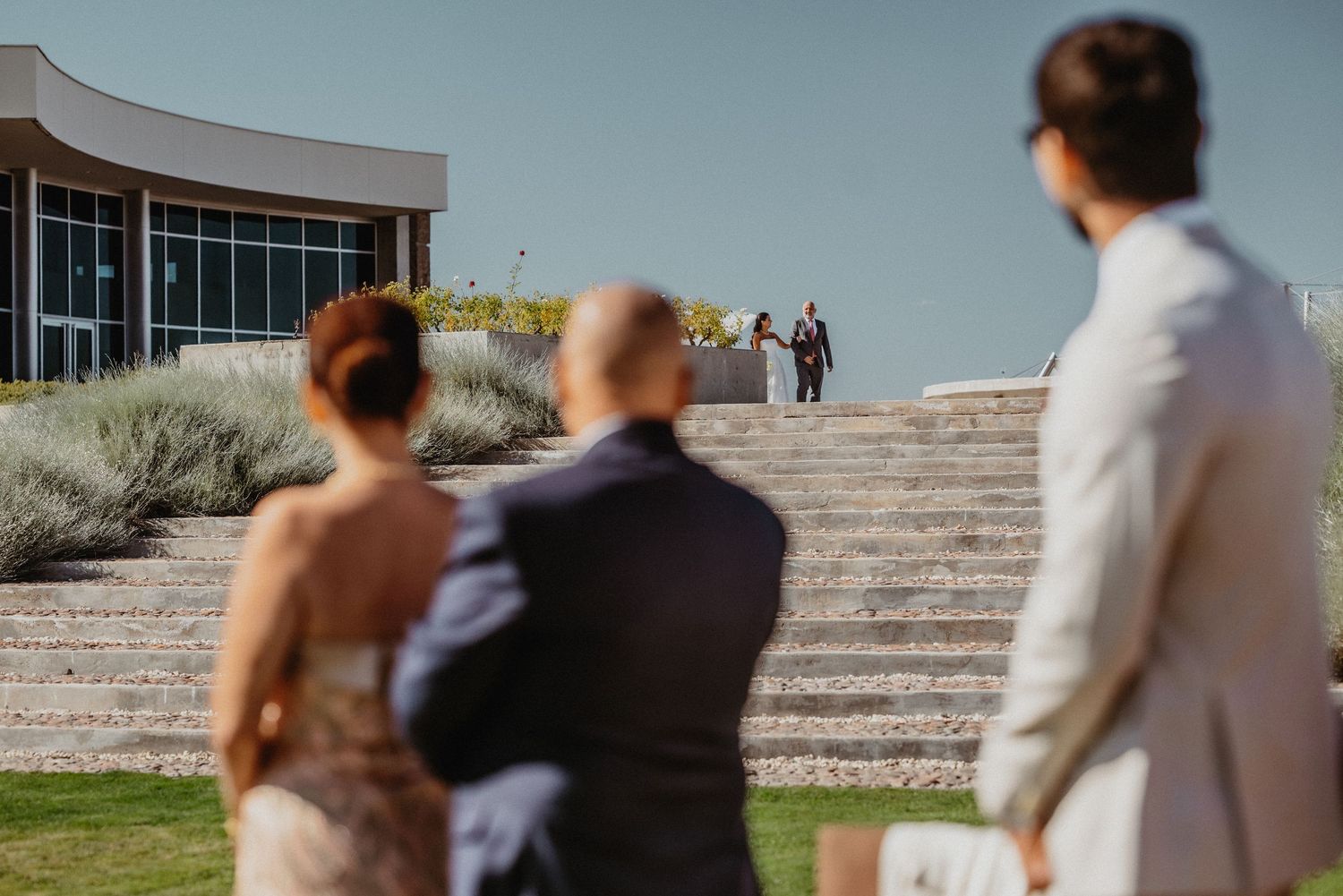 Guests watch as a couple walks down grand stone steps outside a modern venue on a sunny wedding day.
