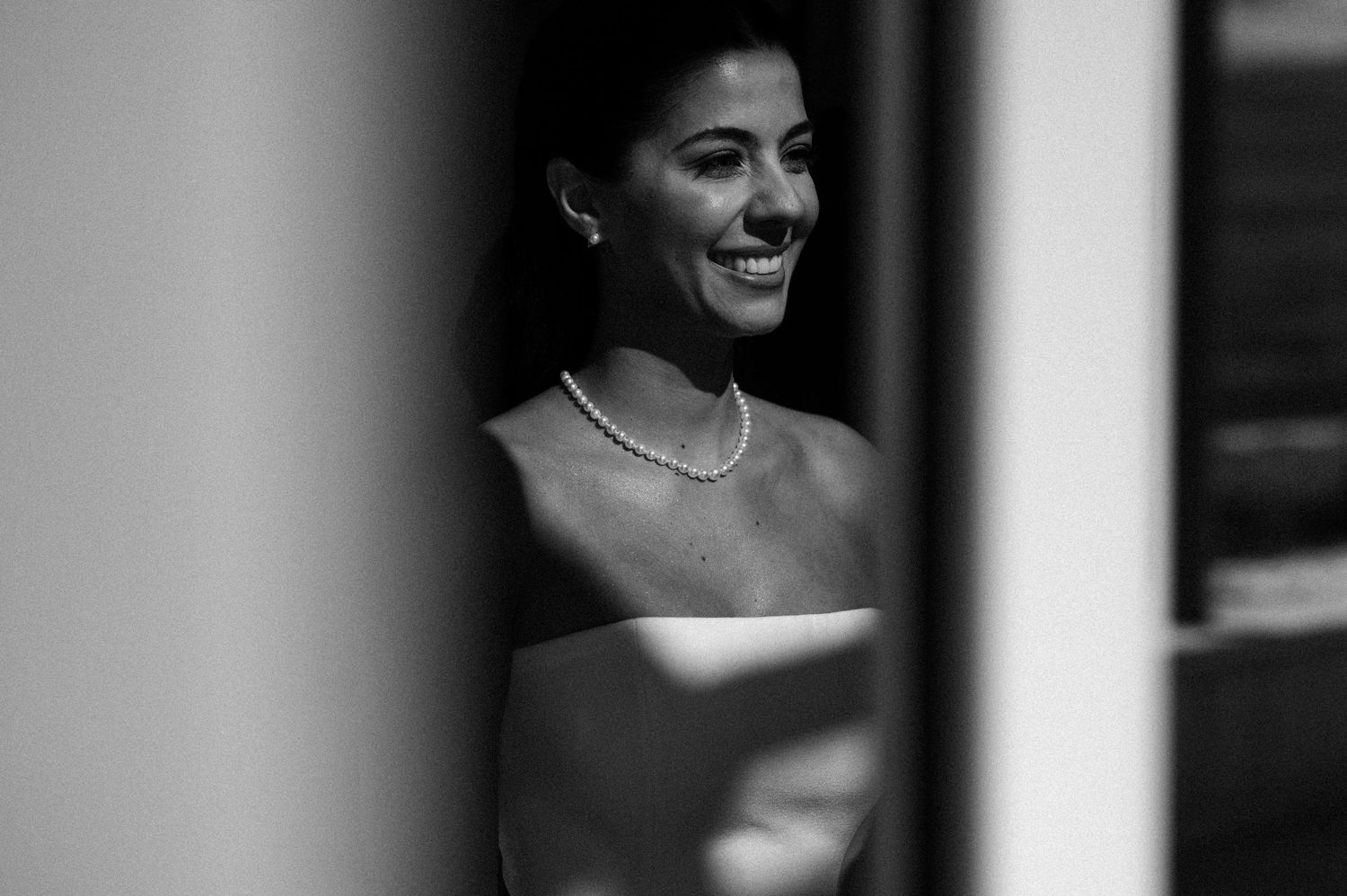 Black and white photo of a smiling bride wearing a pearl necklace and strapless gown, captured through a doorway.