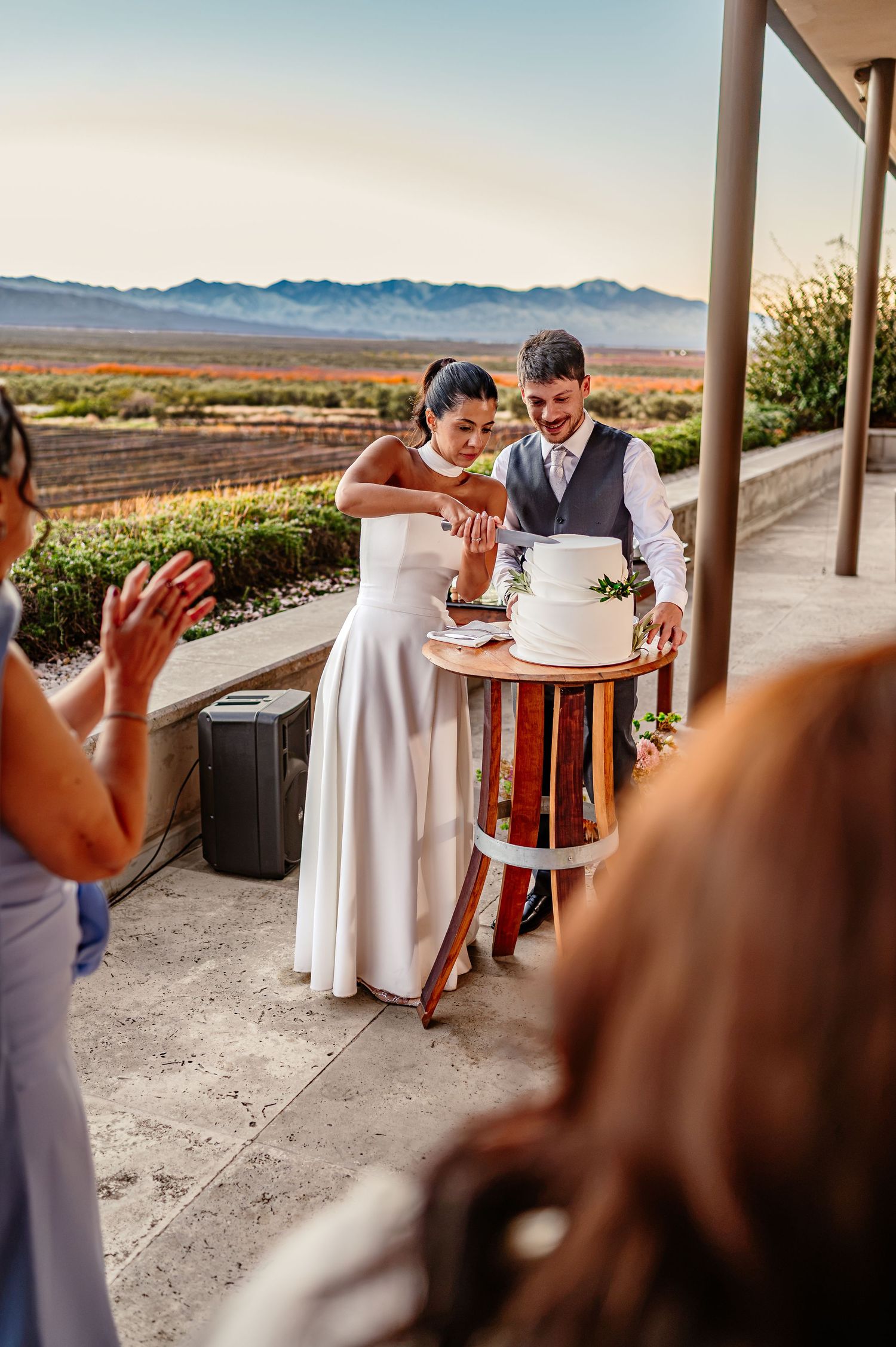Bride and groom cutting white wedding cake outdoors at vineyard venue with mountain views at sunset. fotógrafos de boda en Mendoza