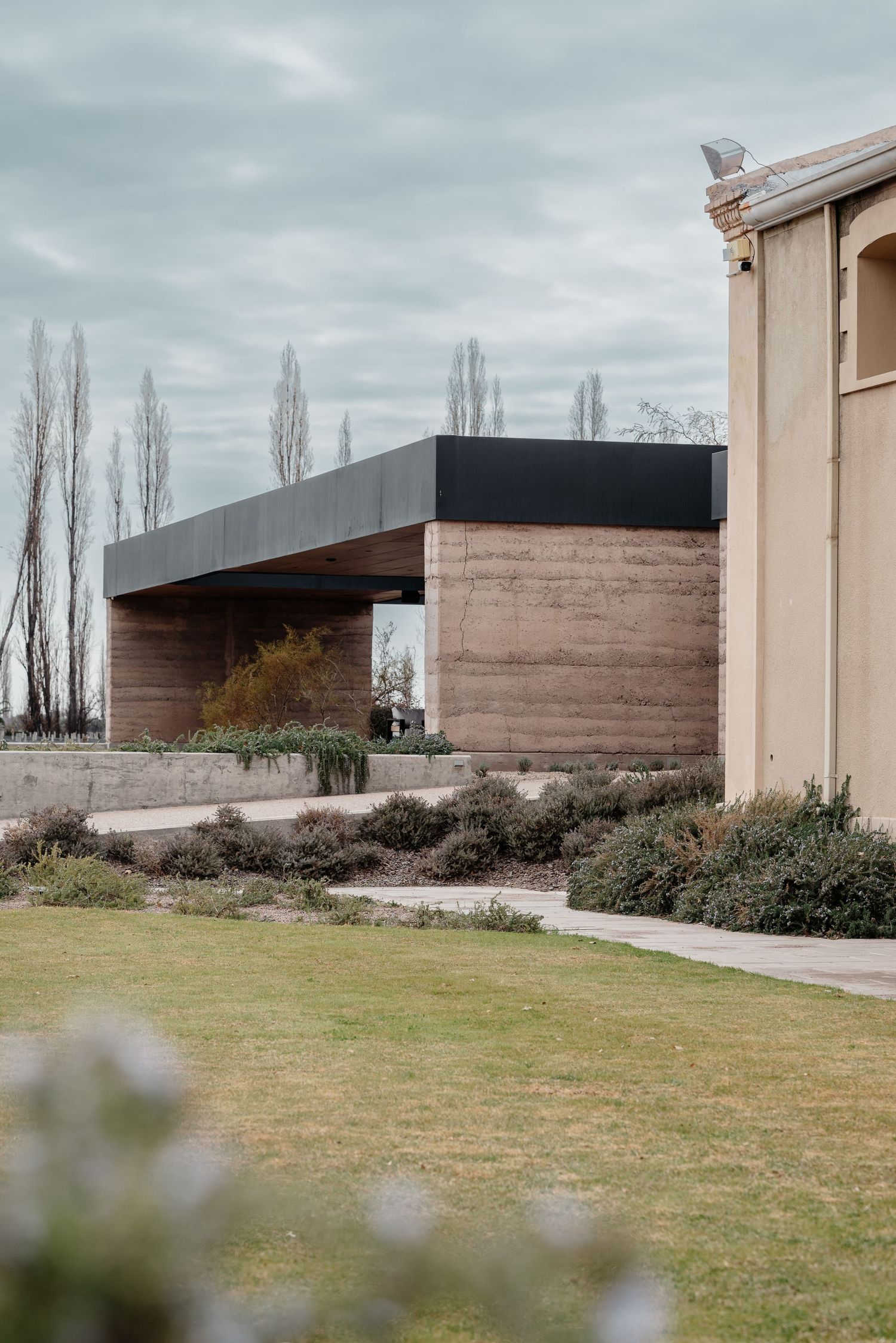 Modern architectural structure with rammed earth walls and black roof beam against a backdrop of tall poplar trees.