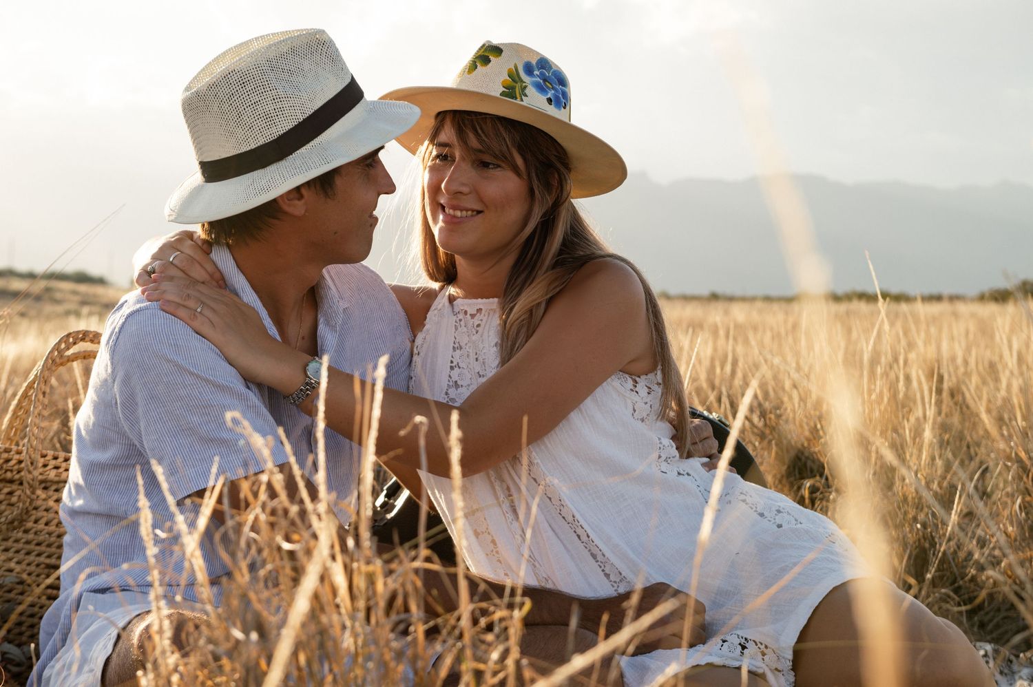Two people in white summer outfits share an intimate moment while sitting in a golden wheat field at sunset.