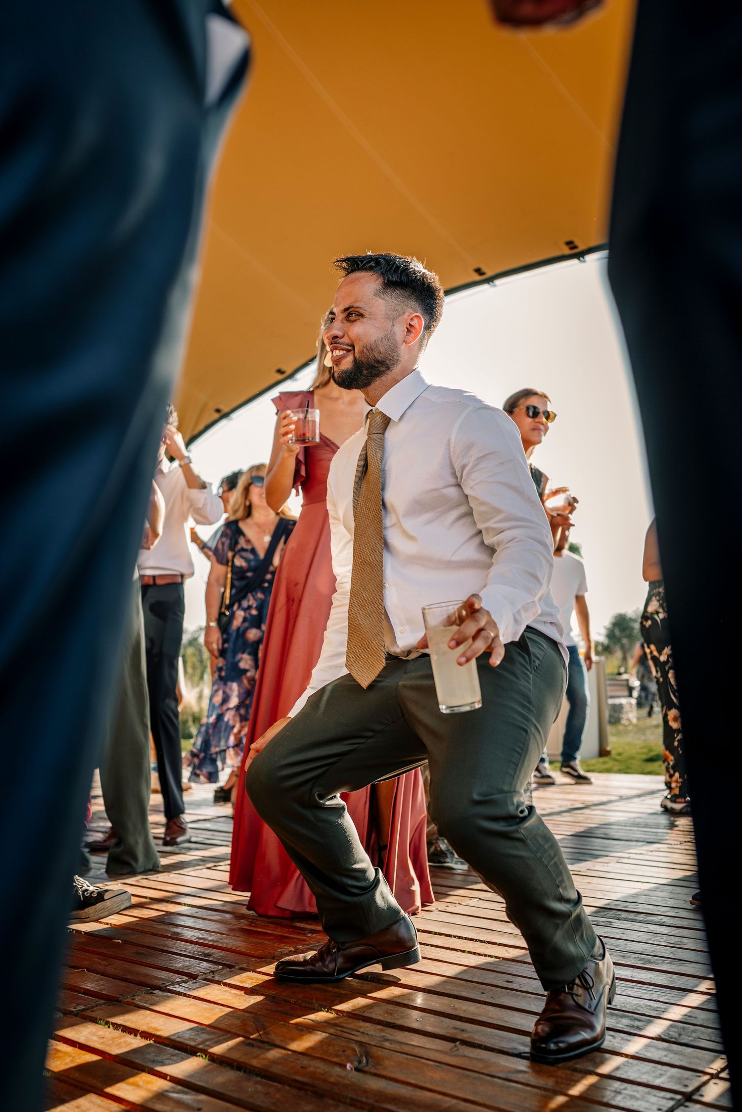 Wedding guests dance energetically in sequence on a wooden deck during sunset at an outdoor reception.
