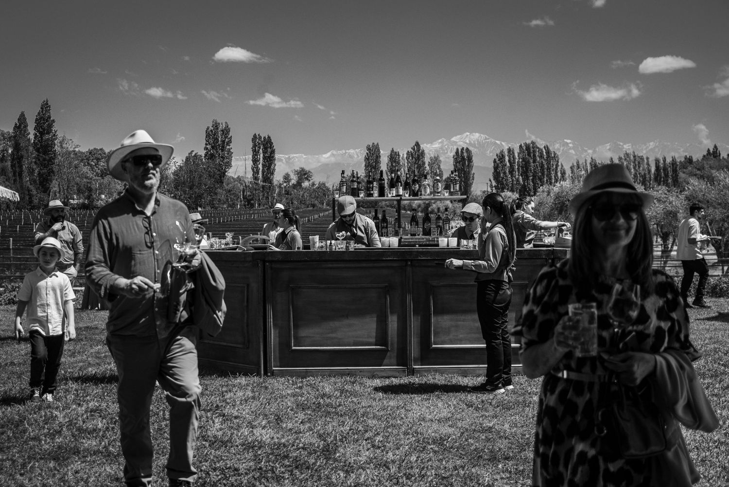 A panoramic black and white landscape view shows people gathered at an outdoor event with tall trees in the background.