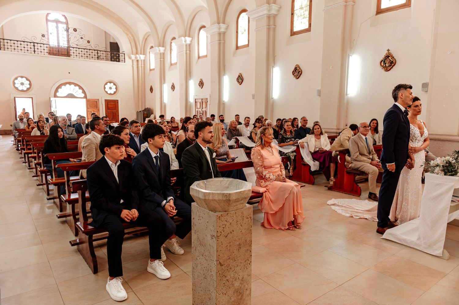 A panoramic view of a traditional church wedding ceremony with guests seated in wooden pews and ornate white walls.