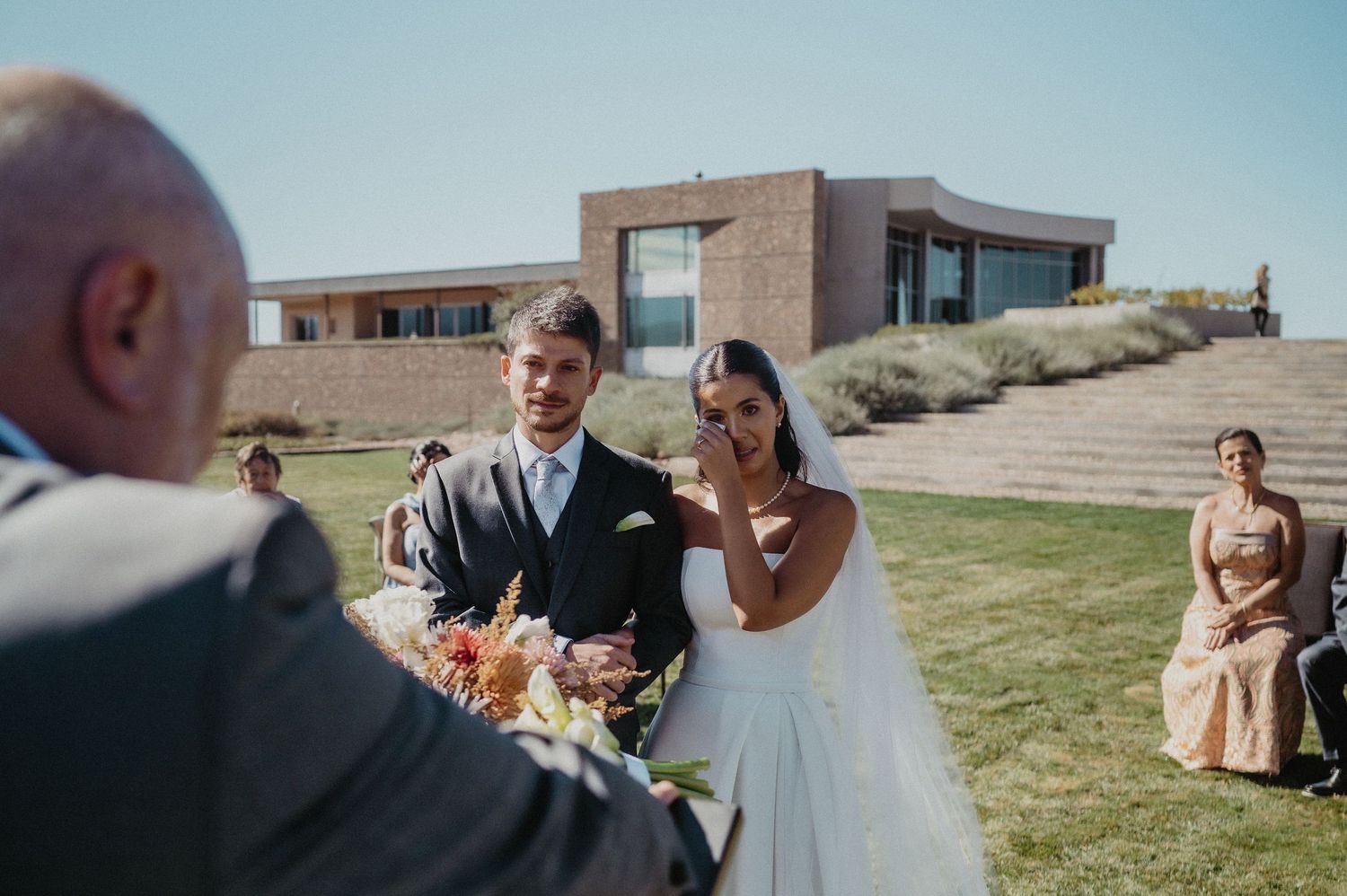 Emotional bride wiping tears during outdoor wedding ceremony with groom and officiant at modern venue.