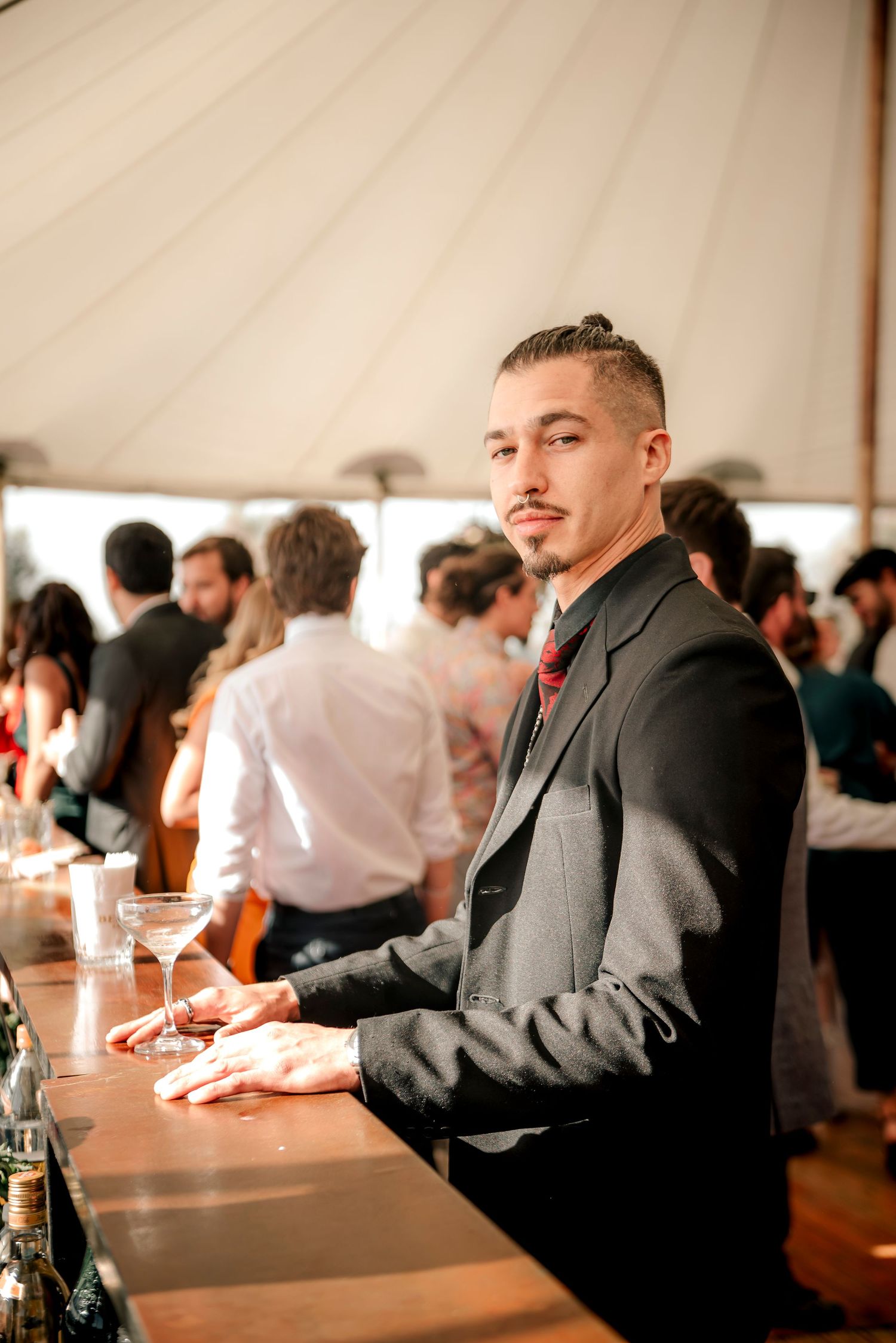 A well-dressed guest in a dark suit jacket stands at a bar during an event under a tent with other attendees behind him.