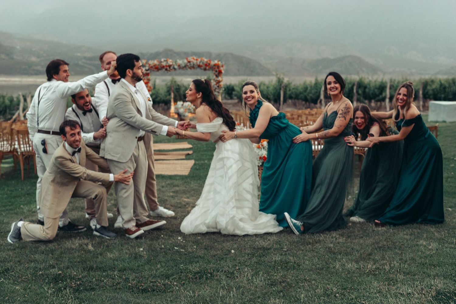 Wedding party sitting in a row on grass having fun pulling each other in playful domino effect pose. fotografos de boda en mendoza