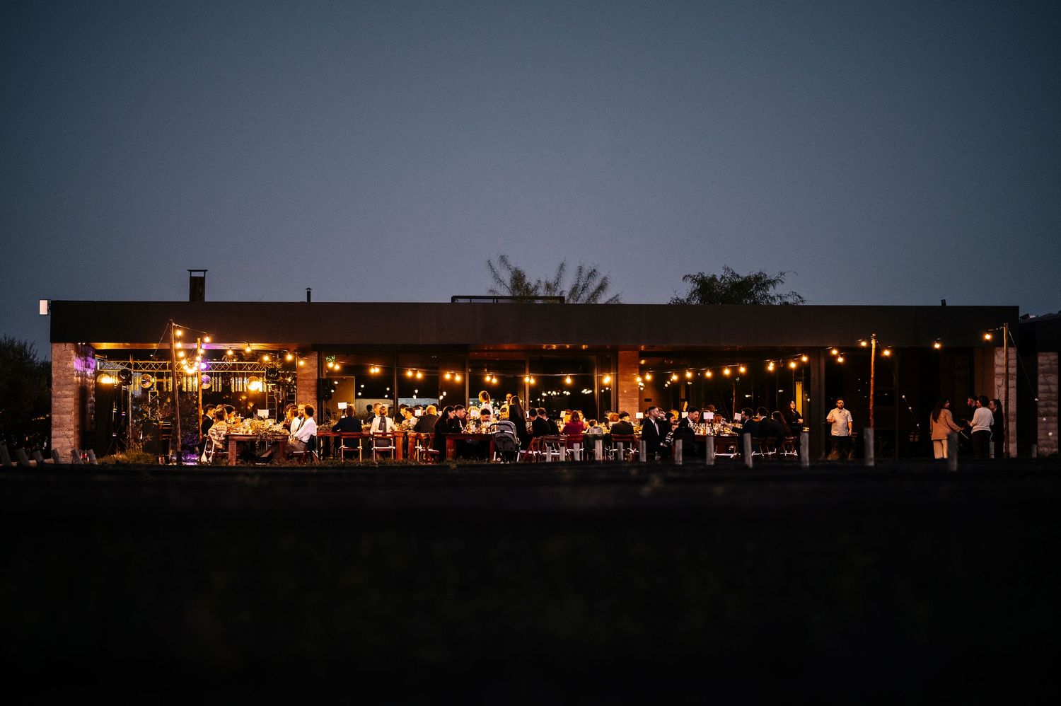 String lights illuminate an outdoor wedding reception venue at dusk creating a warm and romantic atmosphere.