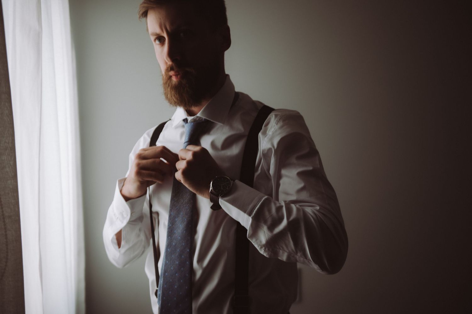 Man in formal attire adjusts tie by window in dramatic side lighting.