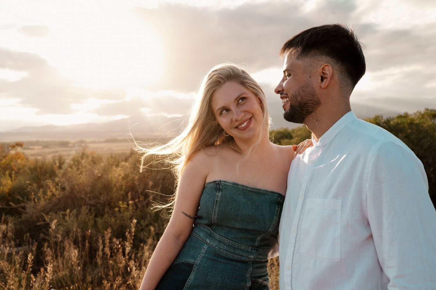 A couple poses together in a field during golden hour as the sun sets behind them in the distance.
