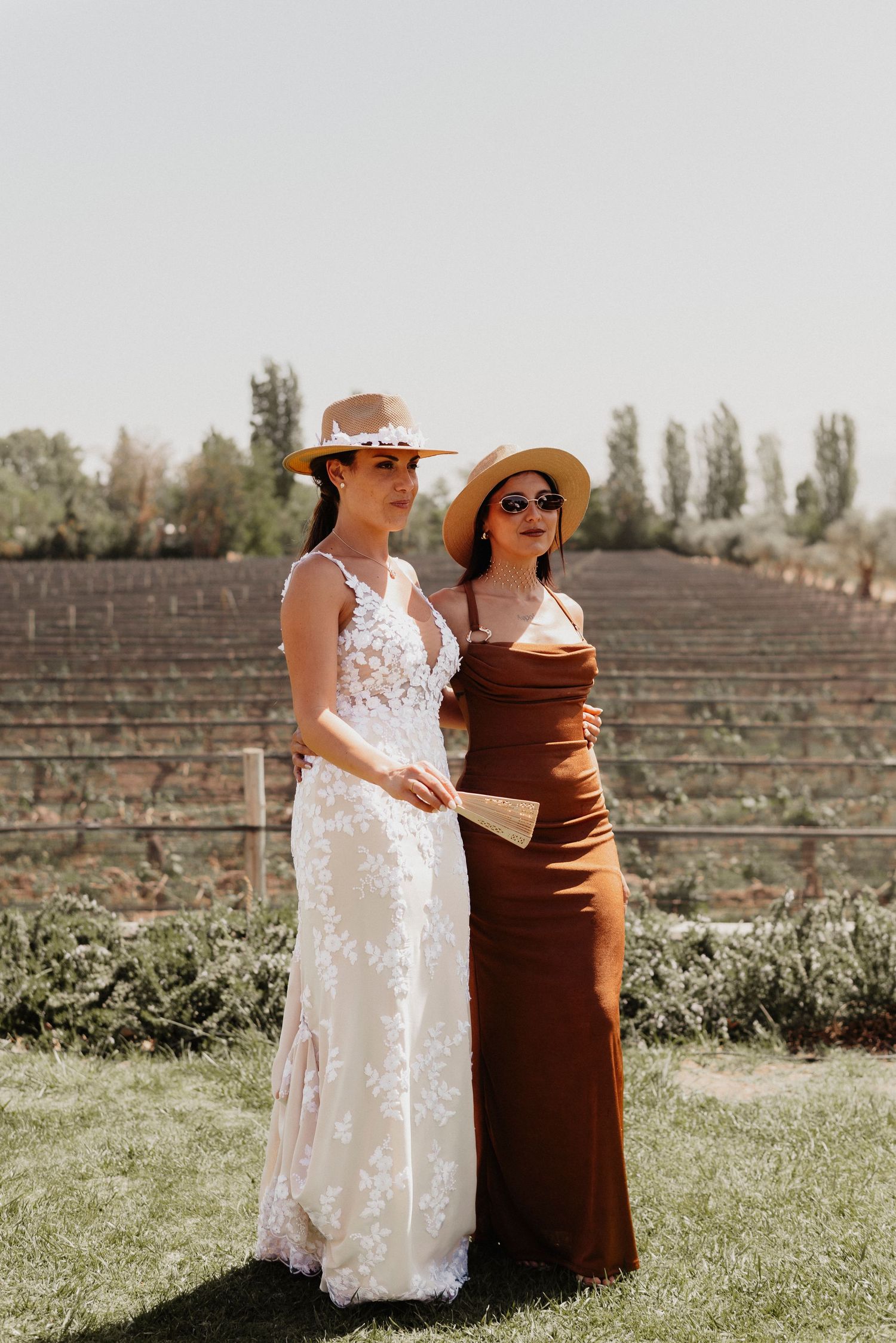 Two people in contrasting white and brown dresses stand together on a grassy lawn with an amphitheater in the background.