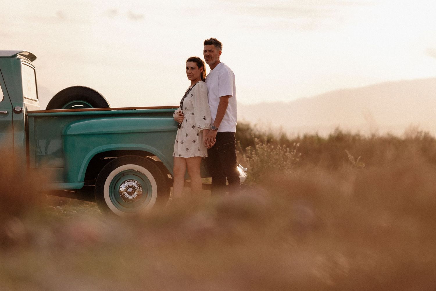 A couple stands beside a vintage turquoise pickup truck in a golden field at sunset.