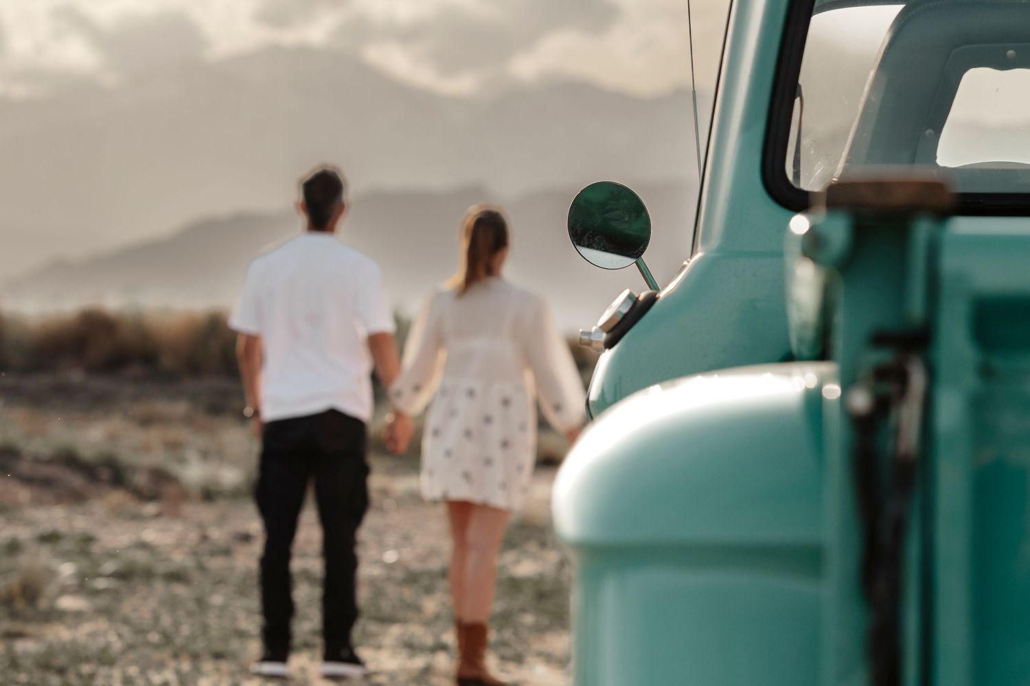A couple holds hands next to a vintage turquoise vehicle in a desert landscape during sunset.