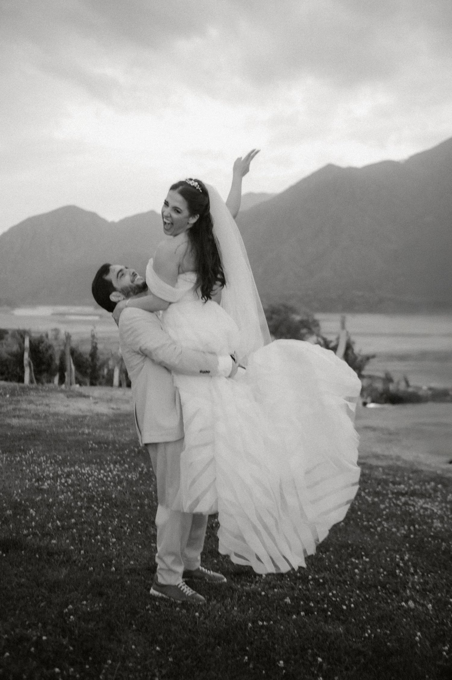 A romantic black and white wedding photo showing a couple embracing outdoors with mountains and a lake in the background.