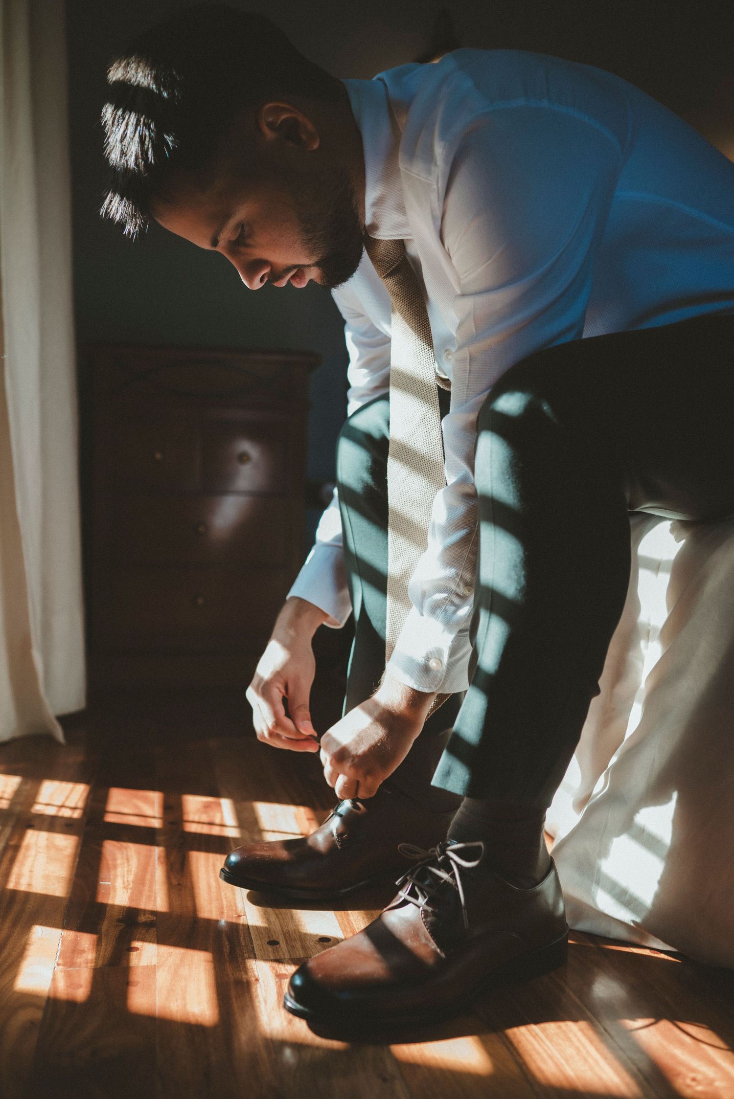 Groom in formal attire adjusts his dress shoes in dramatic window lighting creating shadows on wooden floor.
