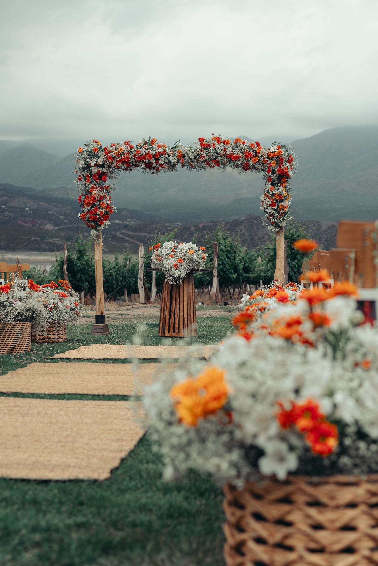 Outdoor wedding ceremony setup with floral arches and mountain backdrop in muted tones and orange flower accents.