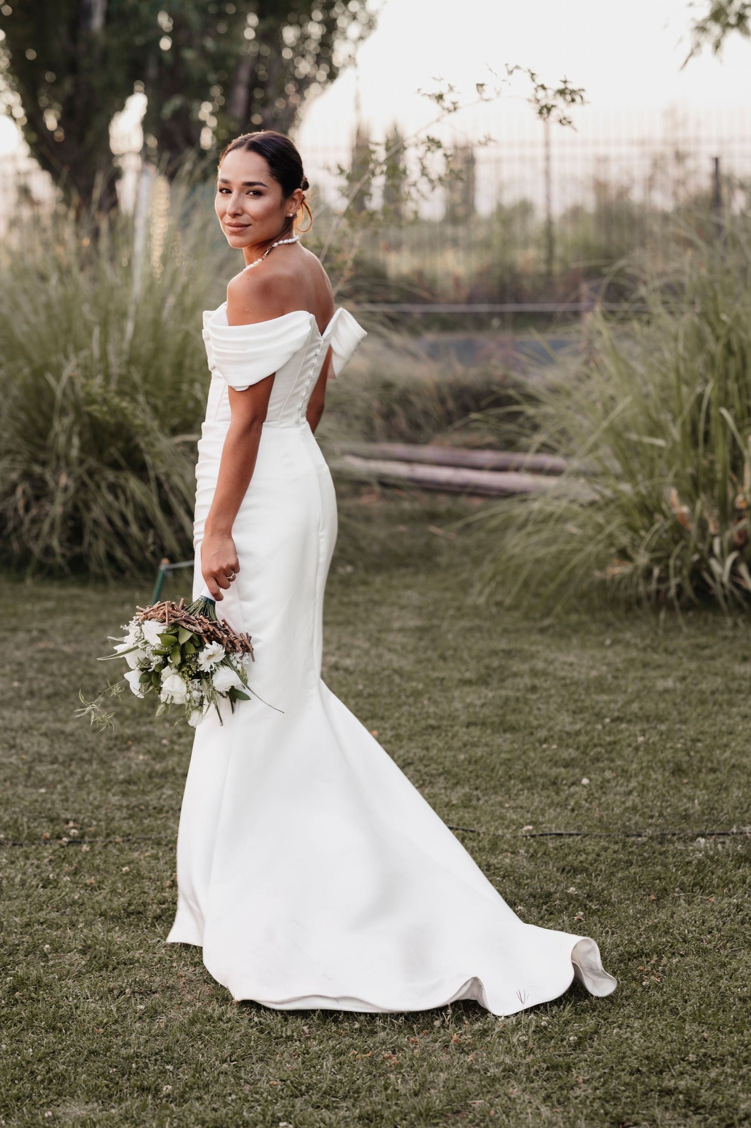 A bride in an elegant white wedding dress poses gracefully on a grassy lawn at sunset.