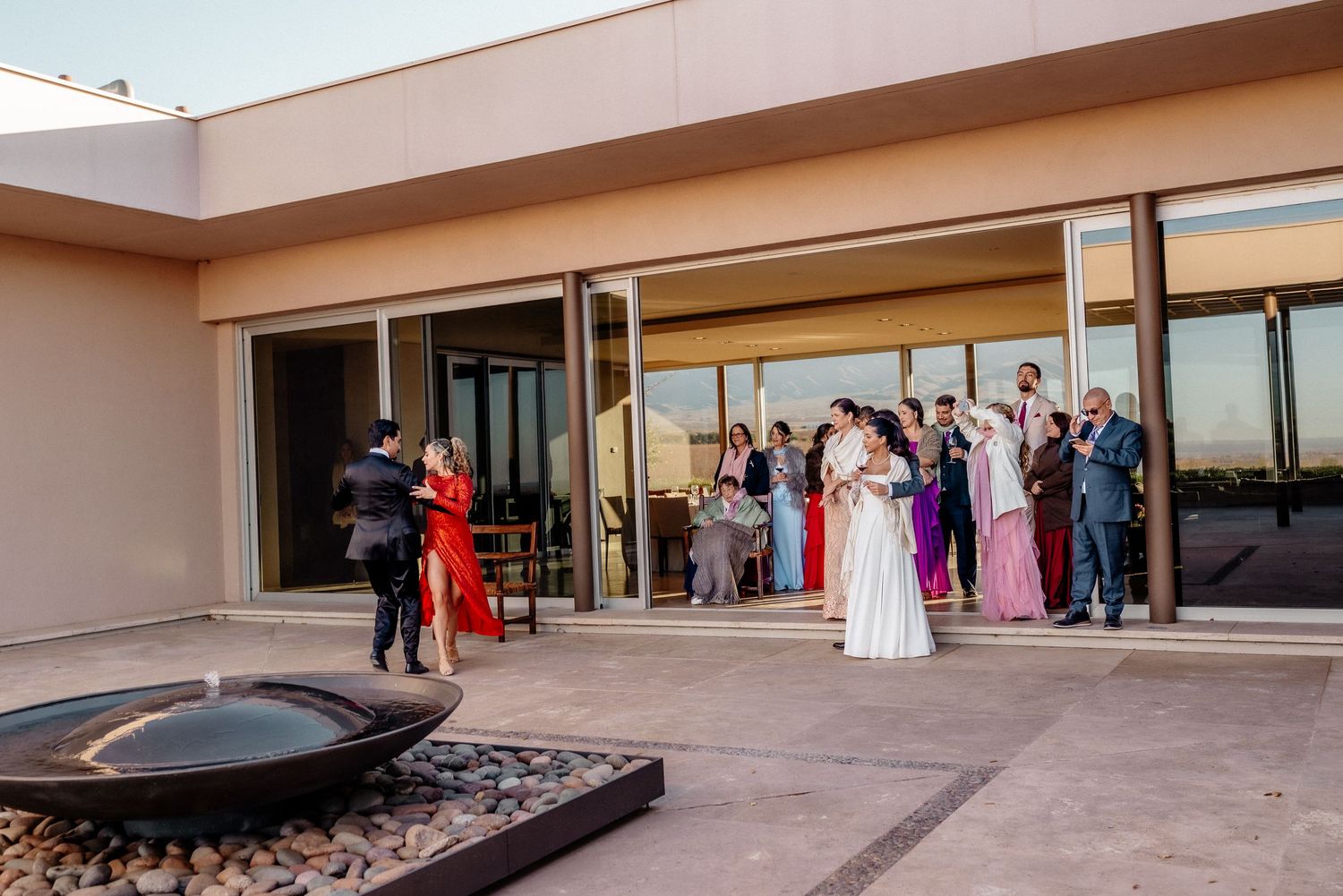 Elegant outdoor wedding reception on a modern patio with guests in formal attire gathered near glass doors at dusk.