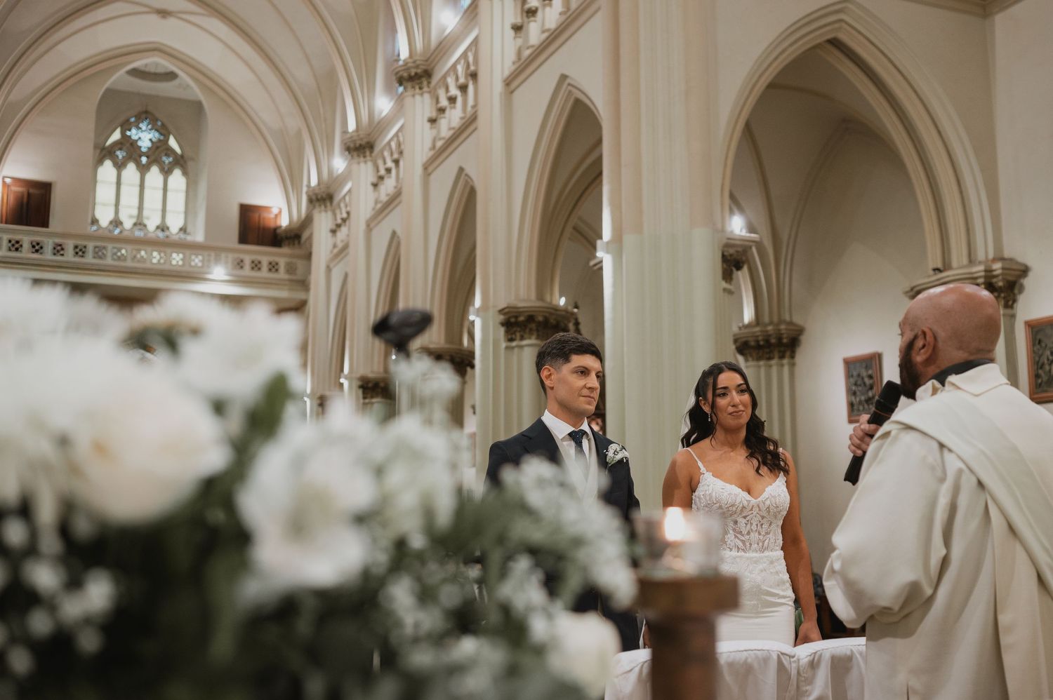 Couple exchanges vows at elegant church wedding ceremony with white floral arrangements and gothic arched ceilings.