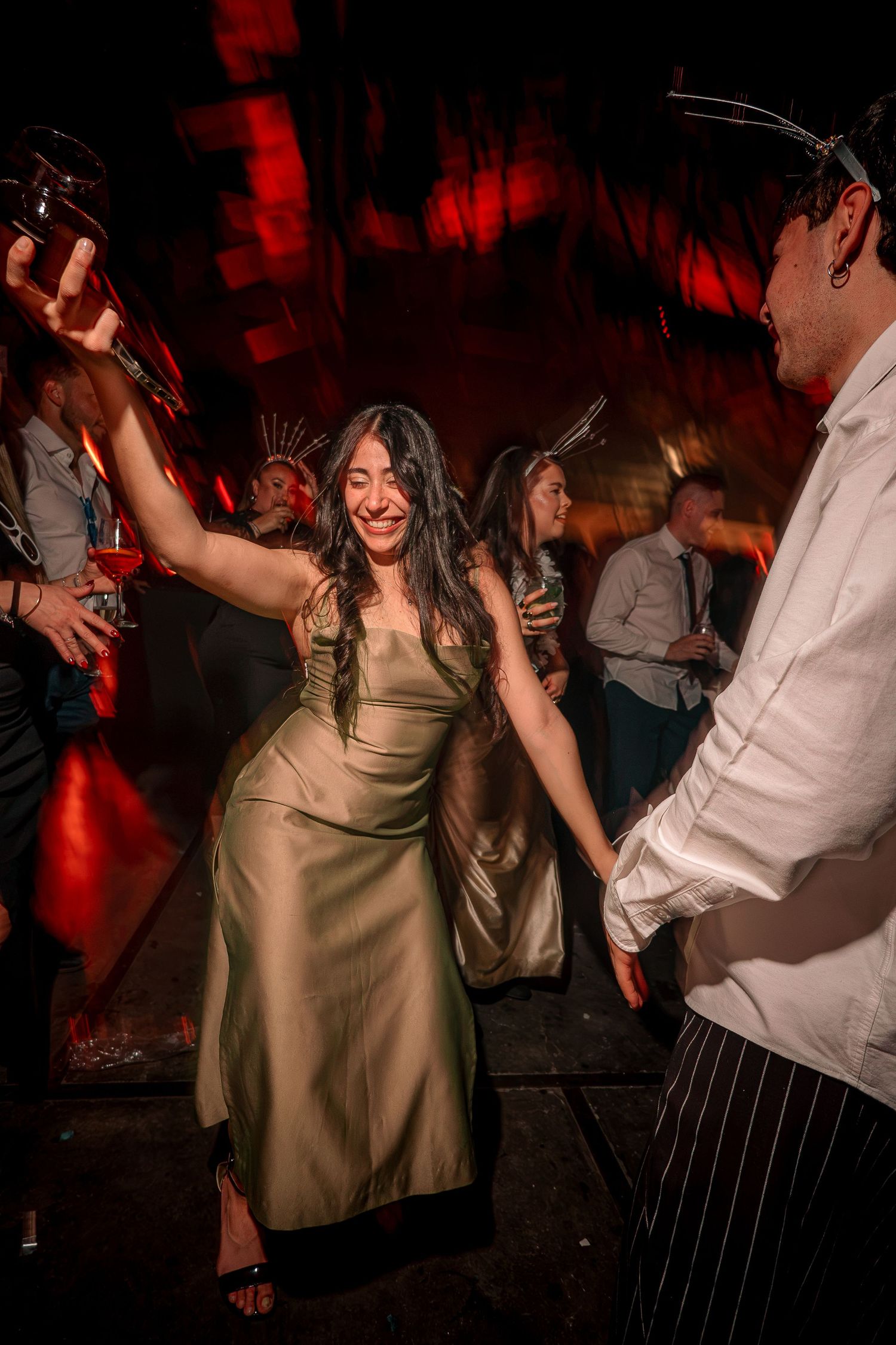 A woman in a gold satin dress dances joyfully at an evening party, holding a drink under dramatic red lighting.