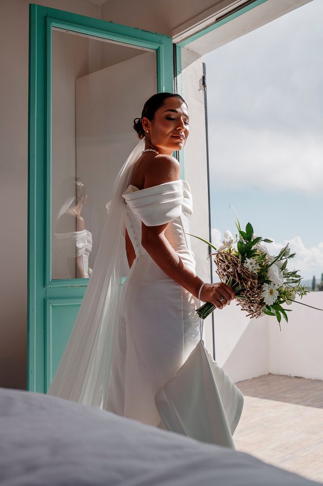 A bride in an elegant white off-shoulder gown poses on a balcony with turquoise accents and holds a natural bouquet. wedding in mendoza