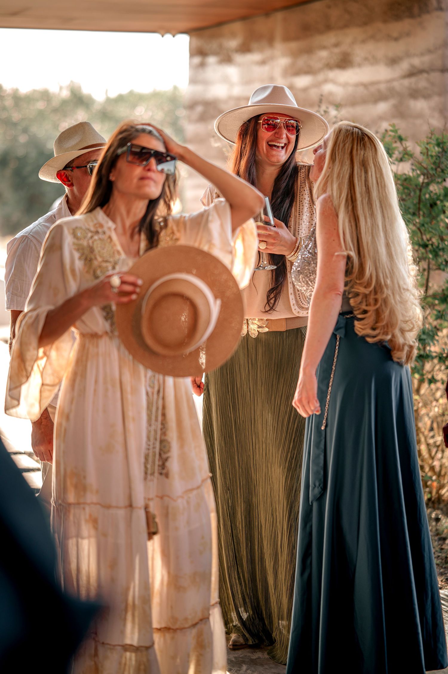 A group of friends in flowy dresses and sun hats share laughter while gathered together outdoors in natural lighting.