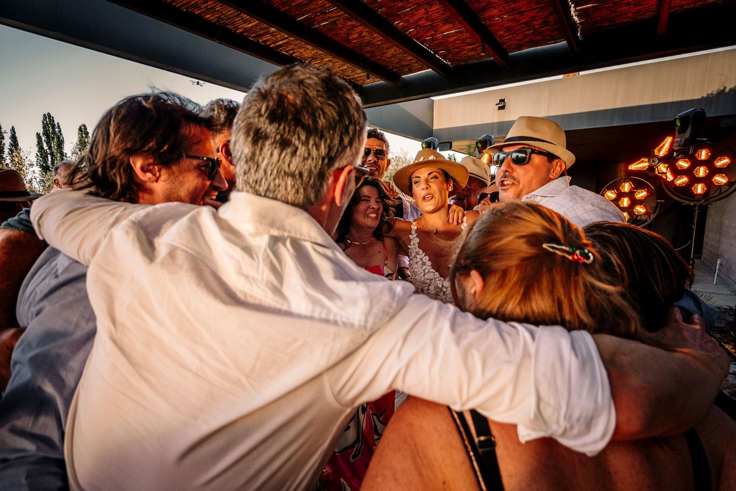 A group of people embrace in a warm hug at a sunset gathering with glowing string lights in the background.