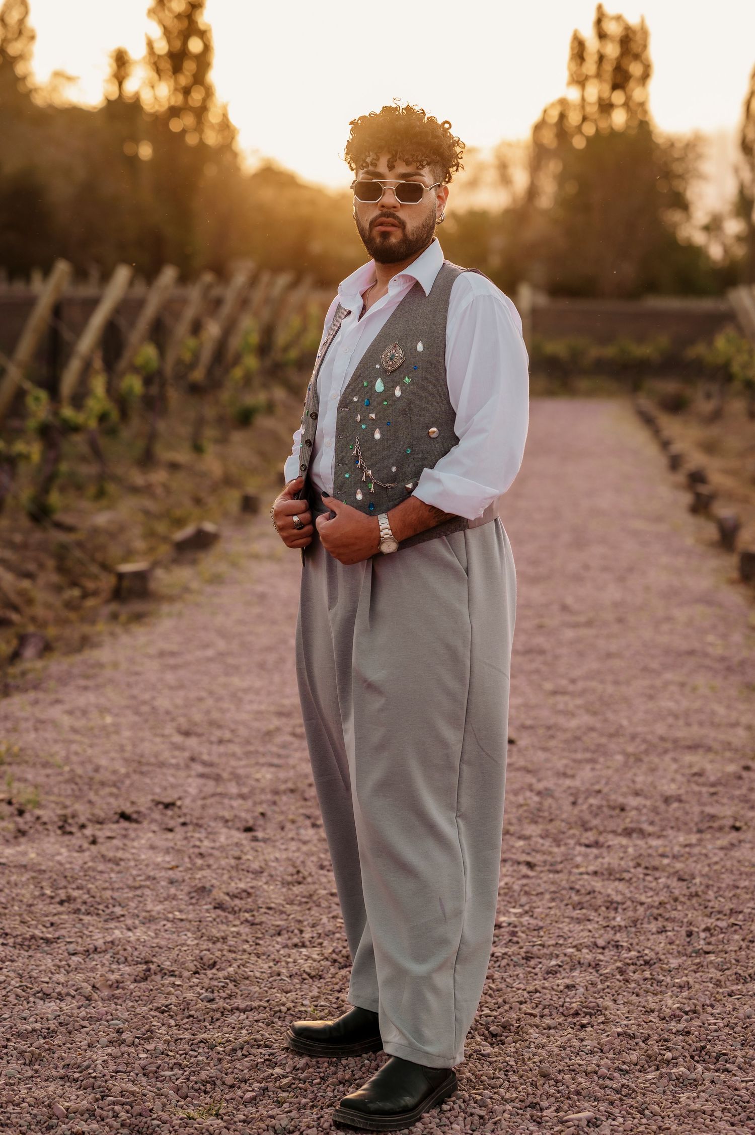 A person in a gray vest and white shirt stands on a gravel path at sunset in a vineyard setting.