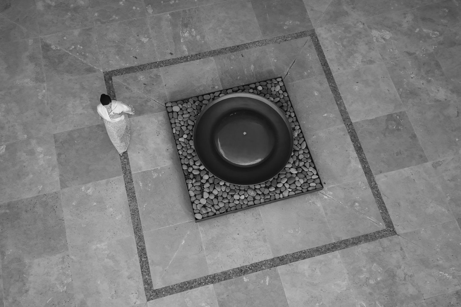 Aerial black and white photo of a person standing beside a circular stone water feature on a tiled floor. fotógrafos de boda en Mendoza