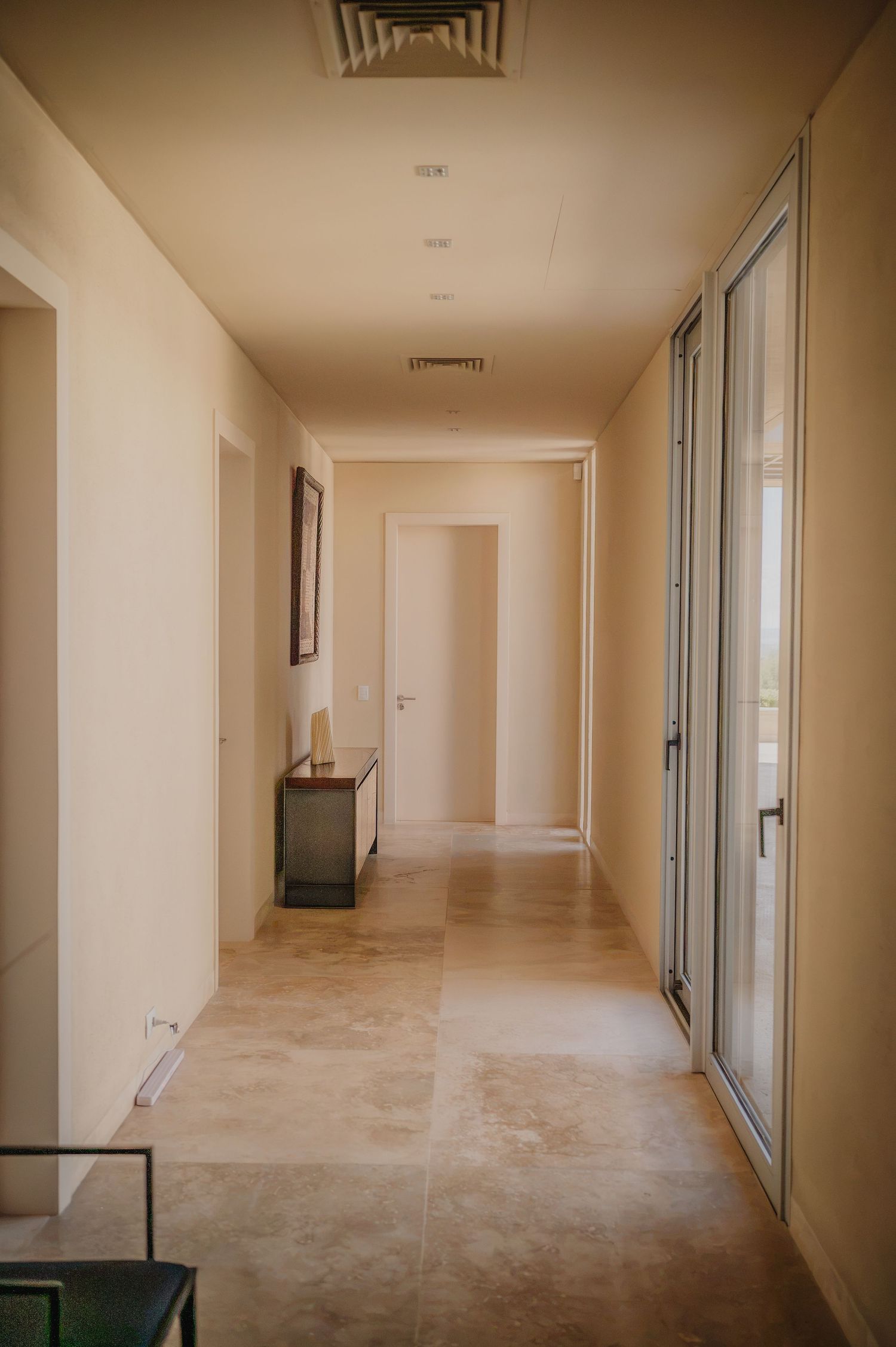 Bright modern hallway with beige tile floors, cream walls, a small console table, and glass sliding doors leading outside.