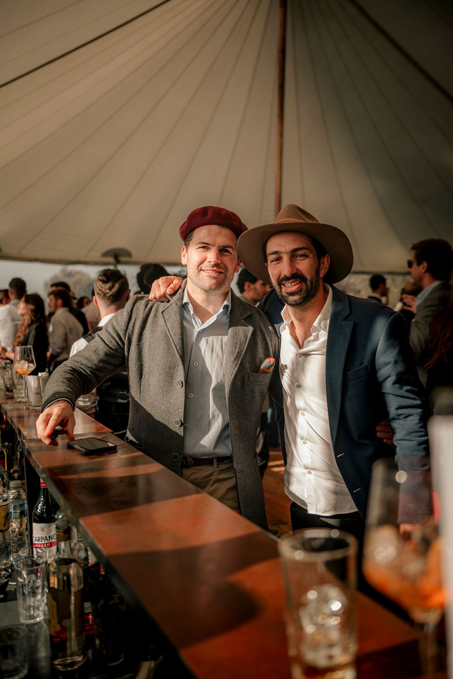 Two friends enjoying drinks together at a rustic bar under a canvas tent with bottles visible on the counter.