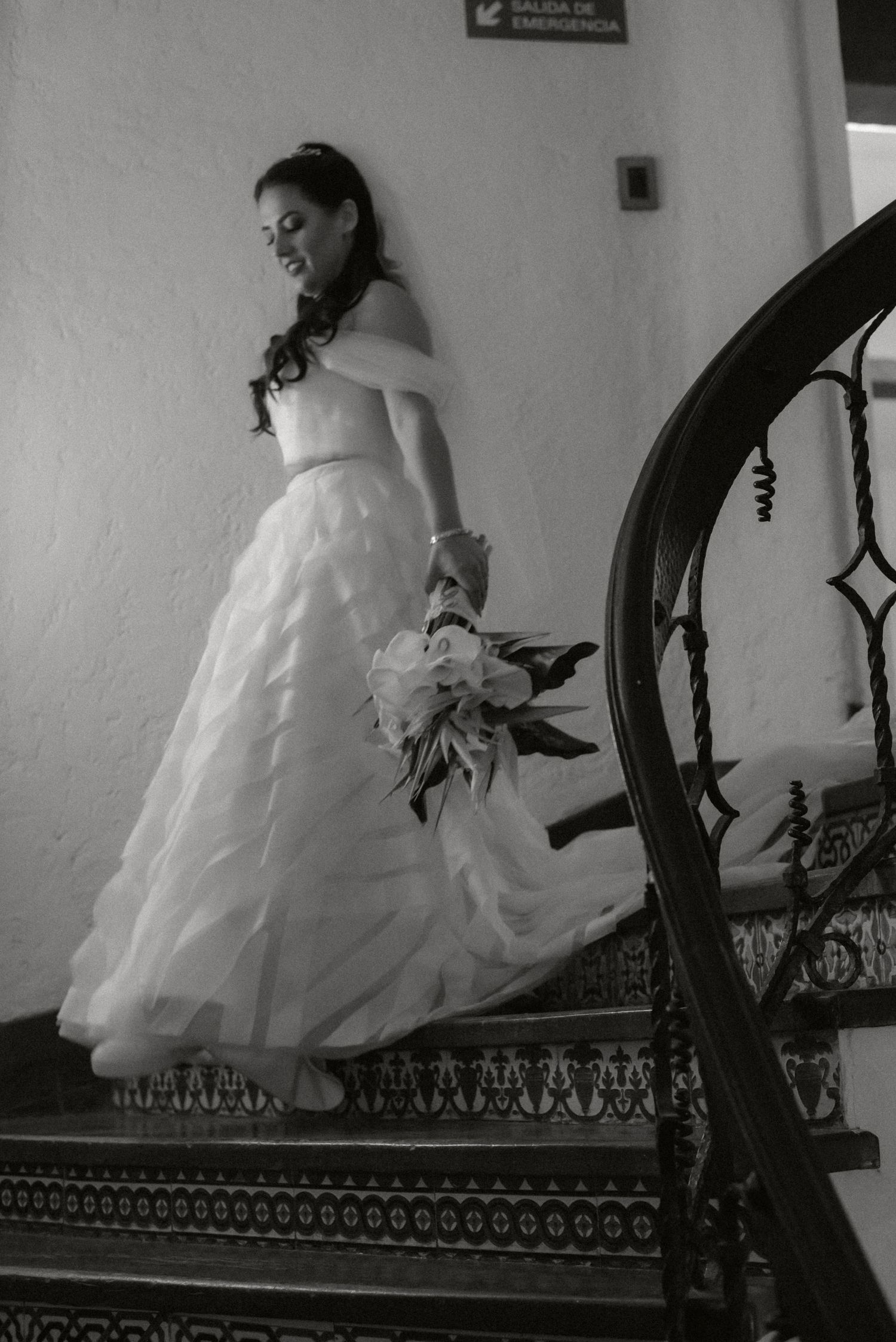 A bride in a flowing white wedding gown sits gracefully on an ornate dark staircase railing in black and white.
