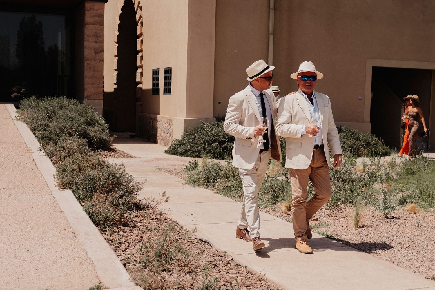 A couple in white and beige attire walks along a beige stucco building exterior during a sunny day.