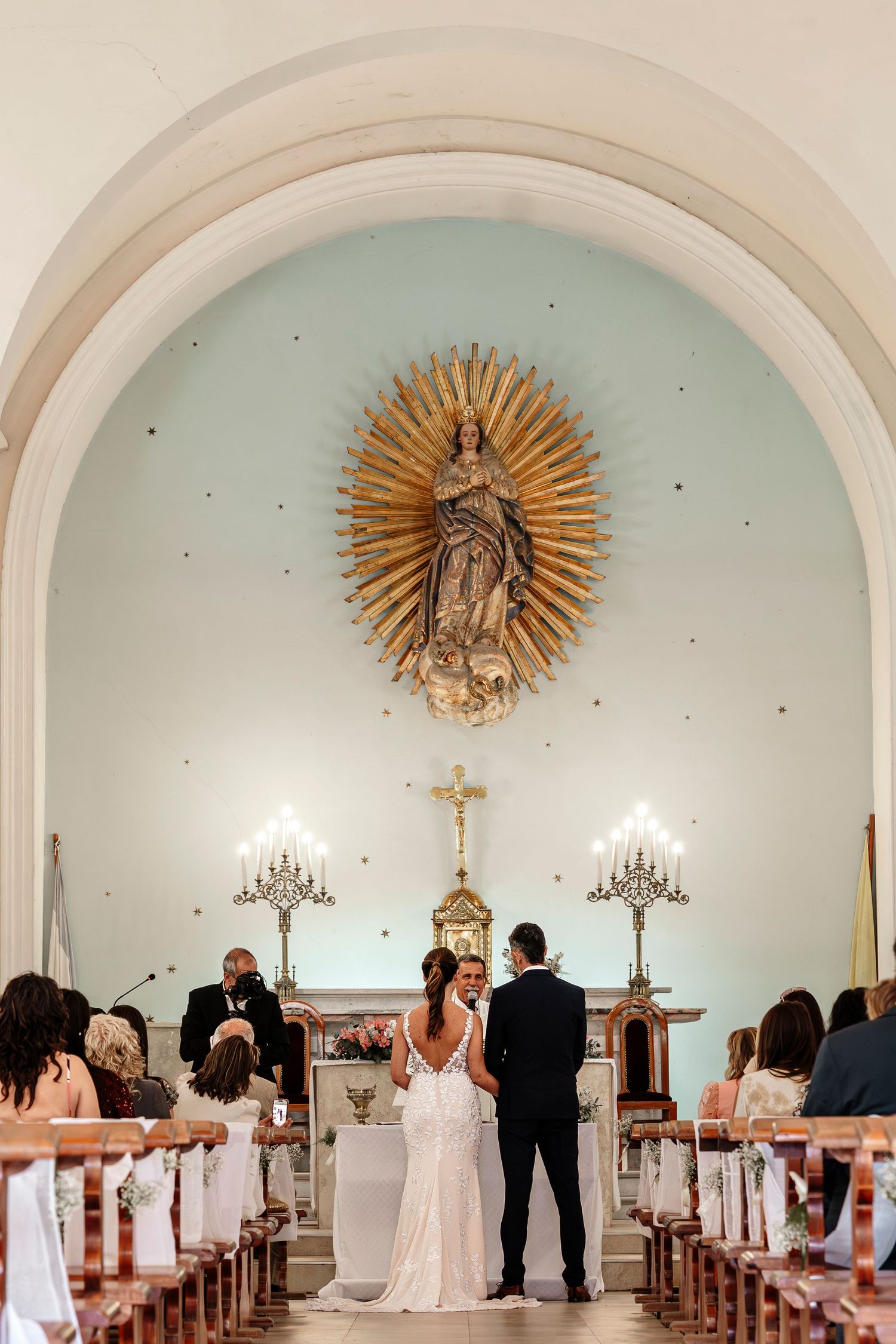 Religious ceremony in a white church interior with ornate golden sunburst decorations and chandeliers.