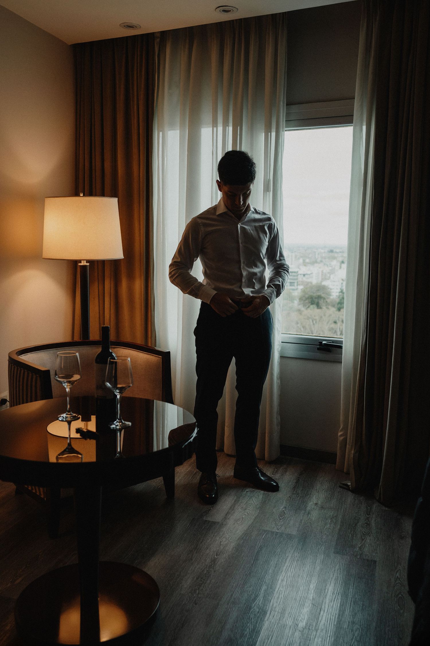 Un hombre con camisa blanca se ajusta los gemelos en una elegante habitación de hotel con cortinas que van del suelo al techo y vino