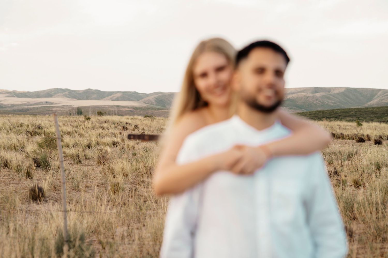 Couple embracing in a scenic field with mountains visible in the distance during golden hour.