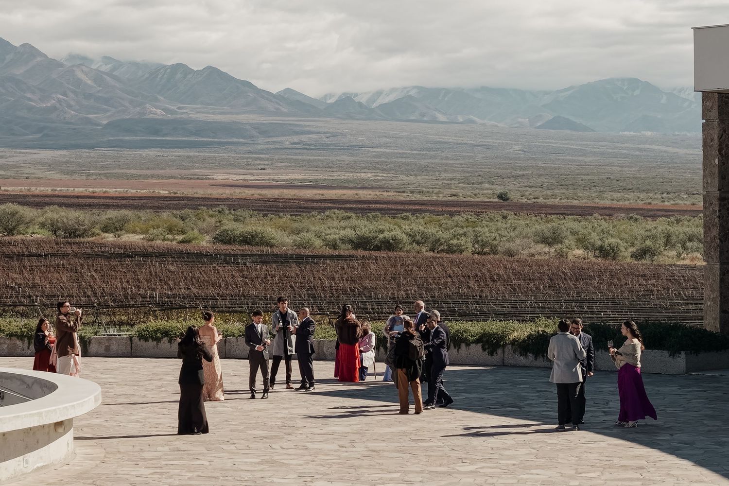 Guests in formal attire mingle on a terrace overlooking vast vineyards and dramatic mountain scenery at an outdoor event.